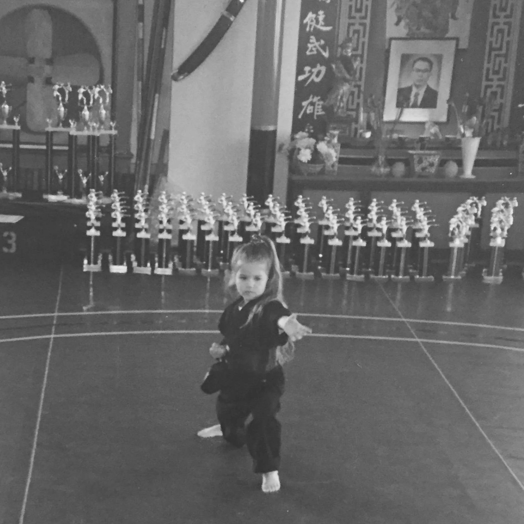 A young girl practicing martial arts in a dojo with a display of trophies and framed photograph of a man in the background.