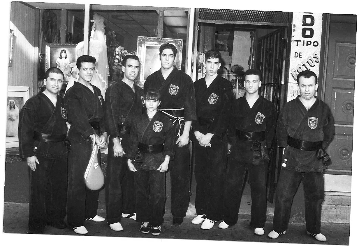 A group of nine people, mostly young men and one young boy, dressed in martial arts uniforms standing in a line in front of a store window displaying paintings and religious figures. They are all wearing black gi uniforms with patches, some with blac