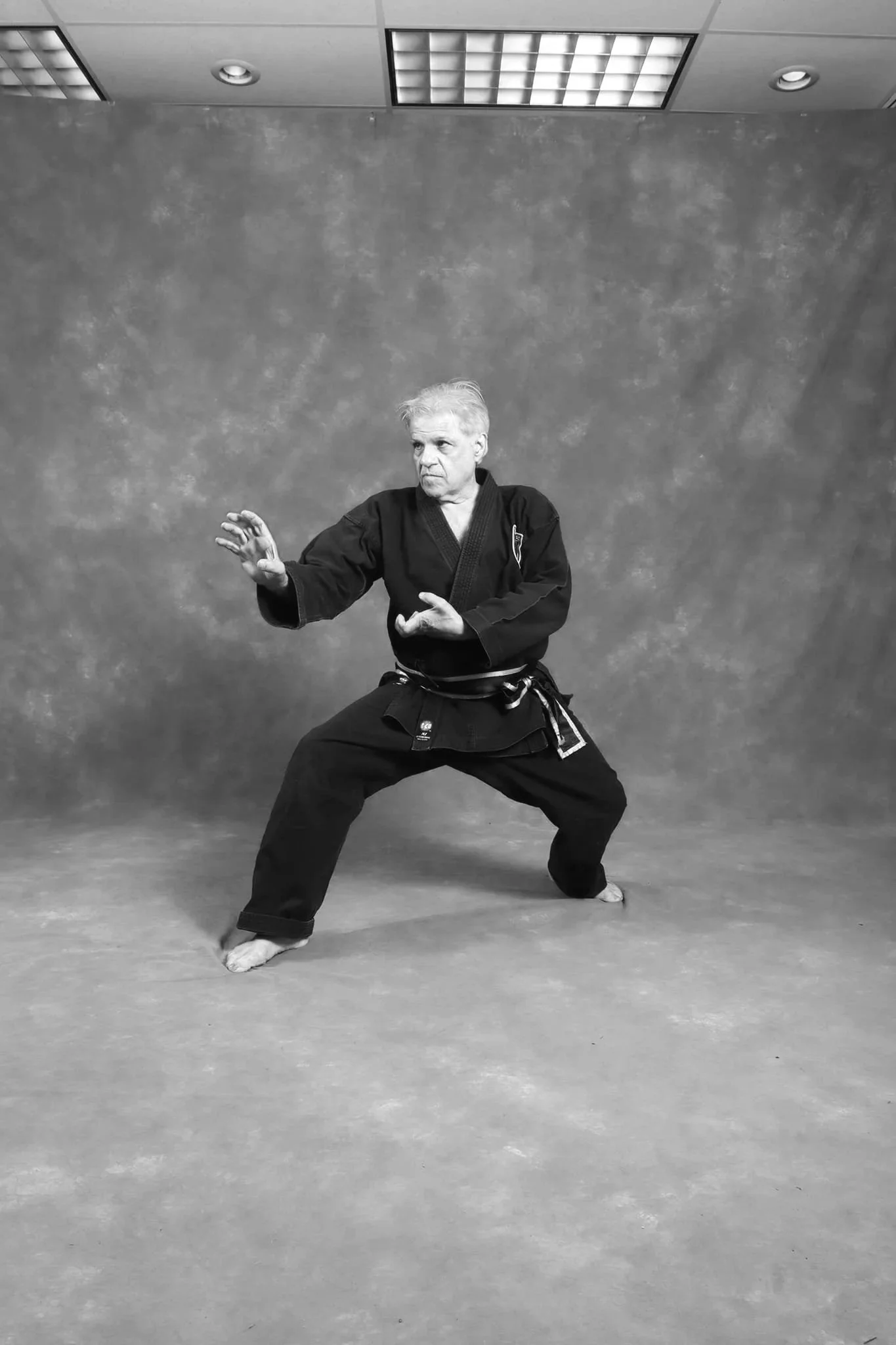 A man dressed in a traditional black martial arts uniform practicing a martial arts stance against a plain backdrop.