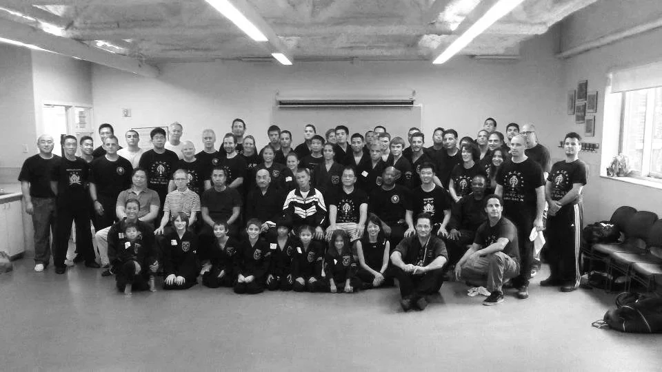 Group of people in a large indoor room, posing for a photo. The group includes children and adults, some wearing matching shirts, with a mix of standing and kneeling positions.