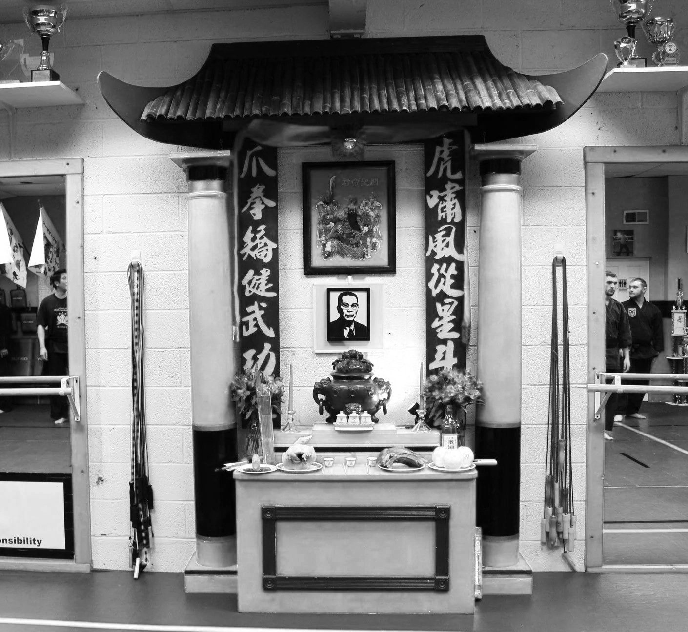 An indoor martial arts altar with traditional Chinese decorations, portraits, and offerings, outside a martial arts training area with two people practicing.