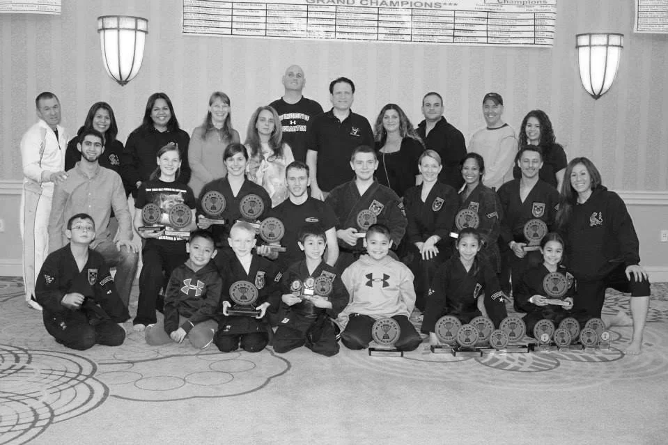 Group photo of children in martial arts uniforms holding trophies and medals, with adults standing behind them, in a decorated indoor setting.