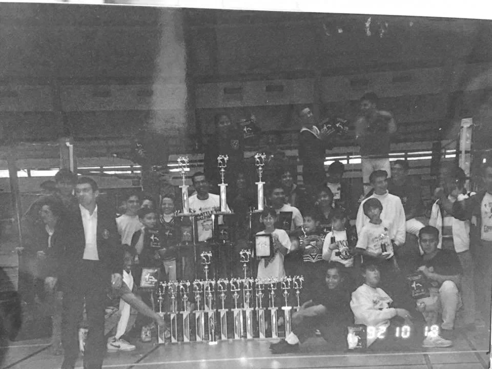 Group of children and adults in a gymnasium with several trophies of different sizes in front of them, celebrating a sports achievement.