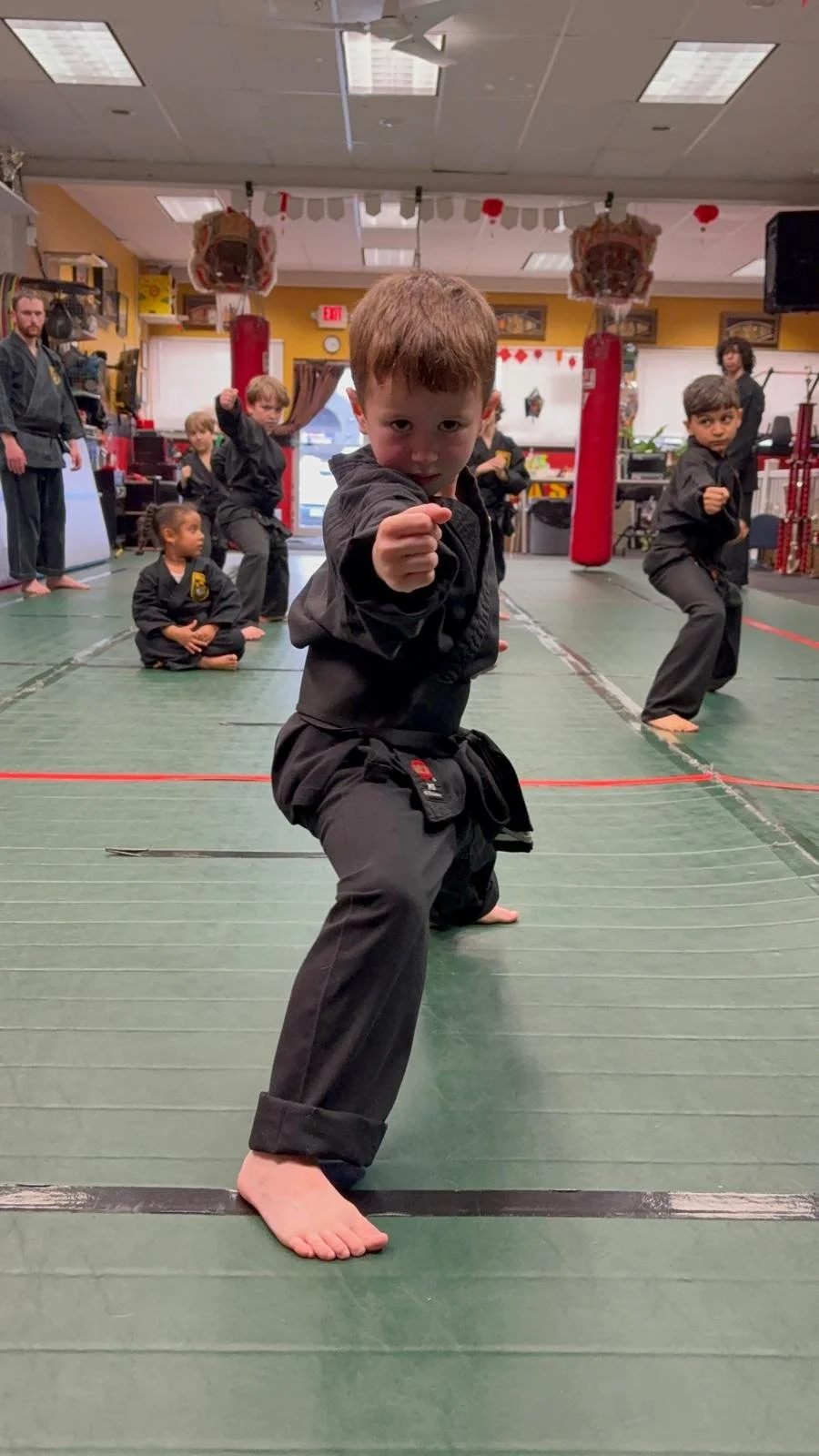 Young children practicing martial arts in a dojo, with one boy in the foreground striking a fighting pose and other children and instructors in the background.