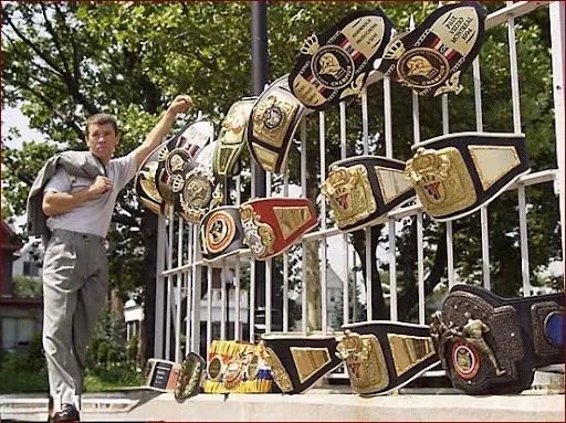 Sifu Vizzio standing next to a display of various (17) championship kickboxing belts on a white metal fence outdoors.