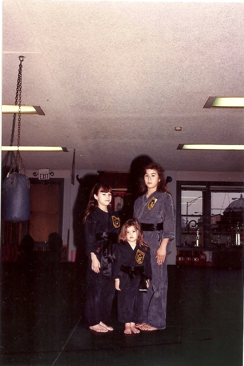 Three girls in martial arts uniforms standing in a dojo, with two of them wearing black traditional uniforms and one girl wearing a gray uniform, all with patches on their uniforms.