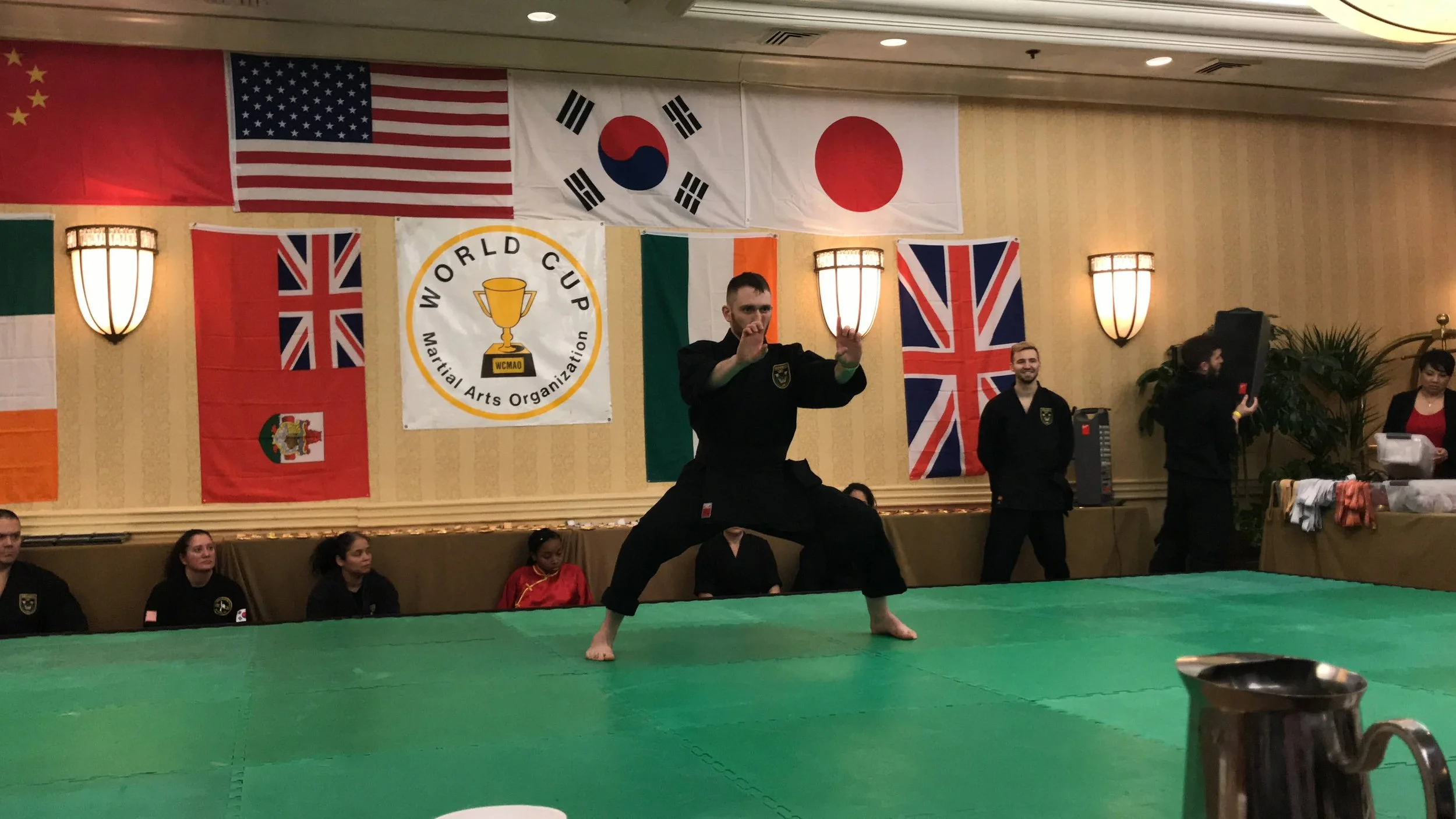 Johnny performing a Martial arts demonstration at a competition or event, with flags of several countries including the United States, South Korea, Japan, China, Ireland, Italy, the United Kingdom, and Ireland displayed on the wall in the background