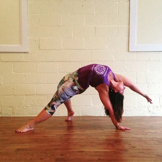 Amanda Kovacs practicing yoga in a side angle pose, with one leg bent and one arm extended overhead, on a wooden floor against a beige brick wall.