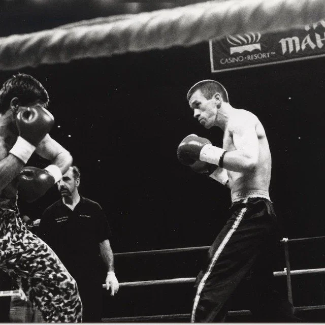 Black and white photo of a boxing match with a boxer in leopard-print shorts and gloves facing an opponent in black pants with a stripe, inside a boxing ring.