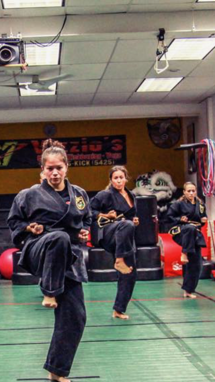 Three Vizzio sisters practicing martial arts in a studio, wearing black uniforms with patches, performing front kicks, with equipment and a banner in the background.