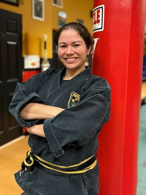 Kitti Vizzio in a black martial arts uniform with a black belt, standing with arms crossed next to a red punching bag, smiling indoors.