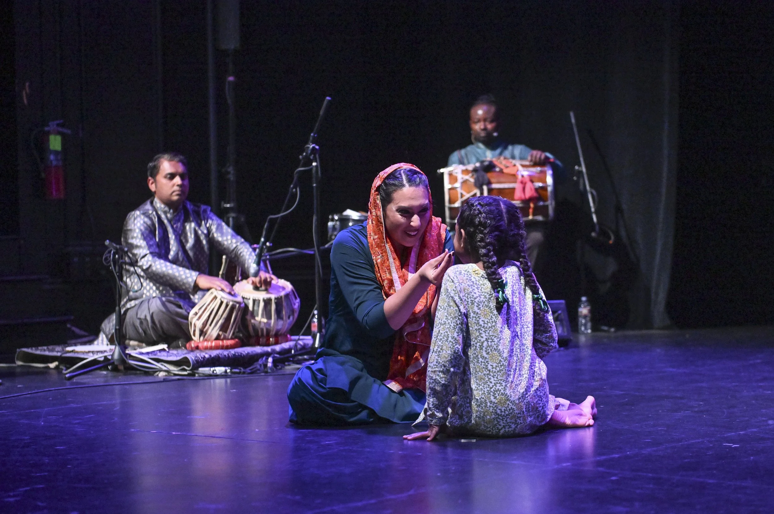 A woman and a girl sitting on the stage, engaging in a conversation, with musicians playing traditional Indian instruments in the background.