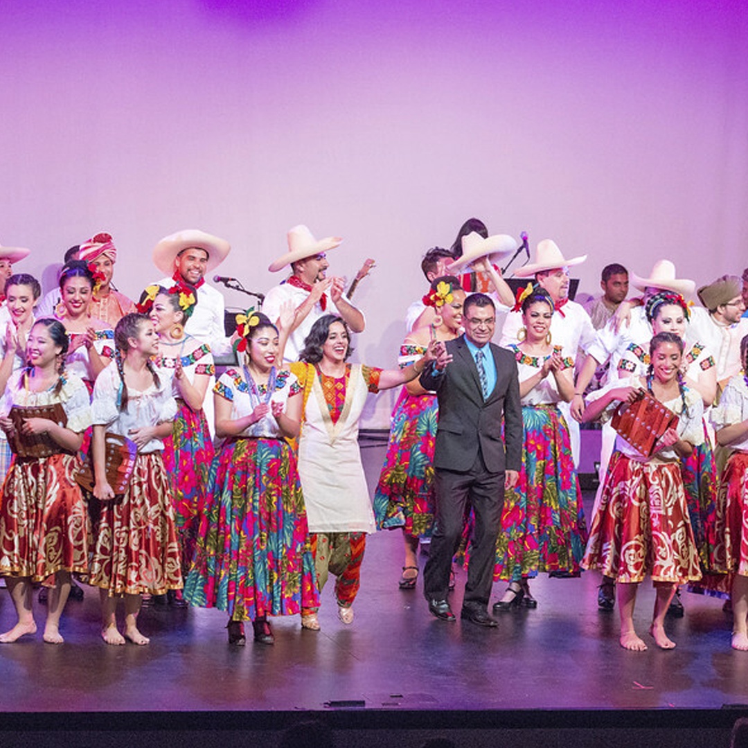 Group of performers in colorful traditional Mexican folk costumes on stage, some wearing sombreros, clapping and smiling after a dance or performance.
