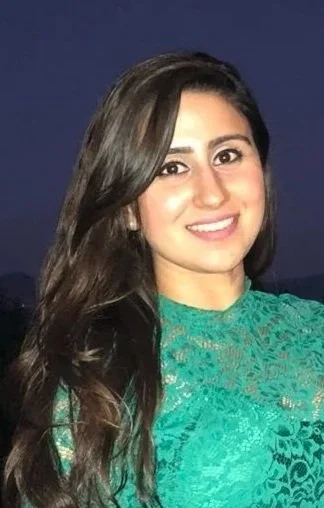 A smiling woman with long, dark hair wearing a teal lace top, standing outdoors during twilight.