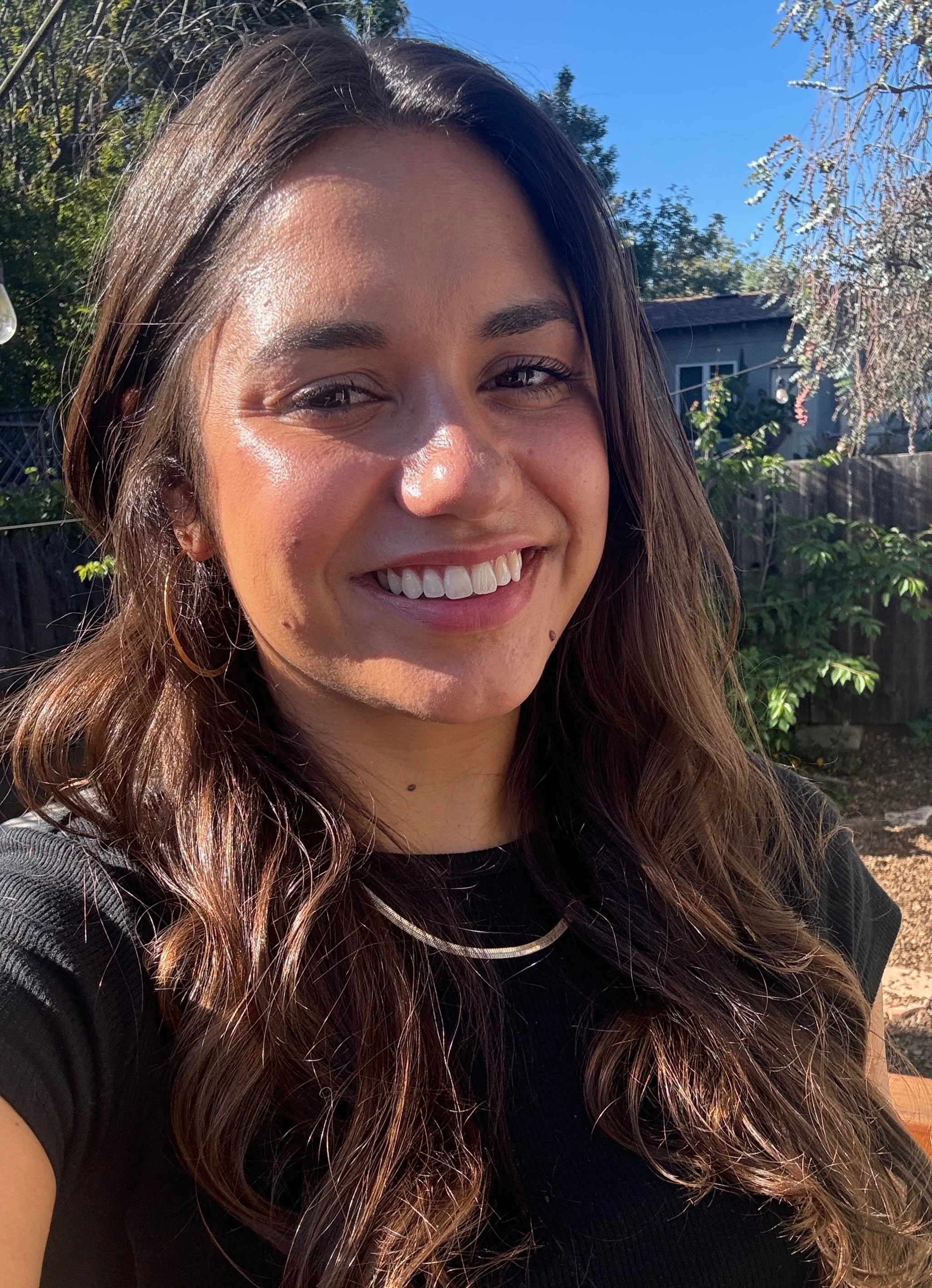A woman smiling outdoors on a sunny day, with trees and a house in the background.