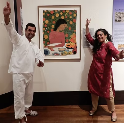 Two people standing in an art gallery, a man on the left in white clothing and a woman on the right in a red traditional outfit, both smiling and dancing in front of a colorful framed painting of a woman with dark hair resting her head on her hand.