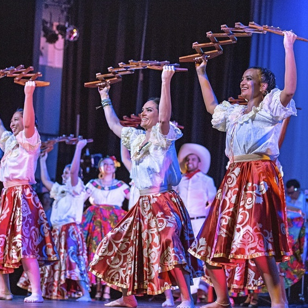 Sweethearts dressed in traditional Mexican attire performing a folkloric dance on stage, holding wooden castanets above their heads, with colorful costumes and a dark background.