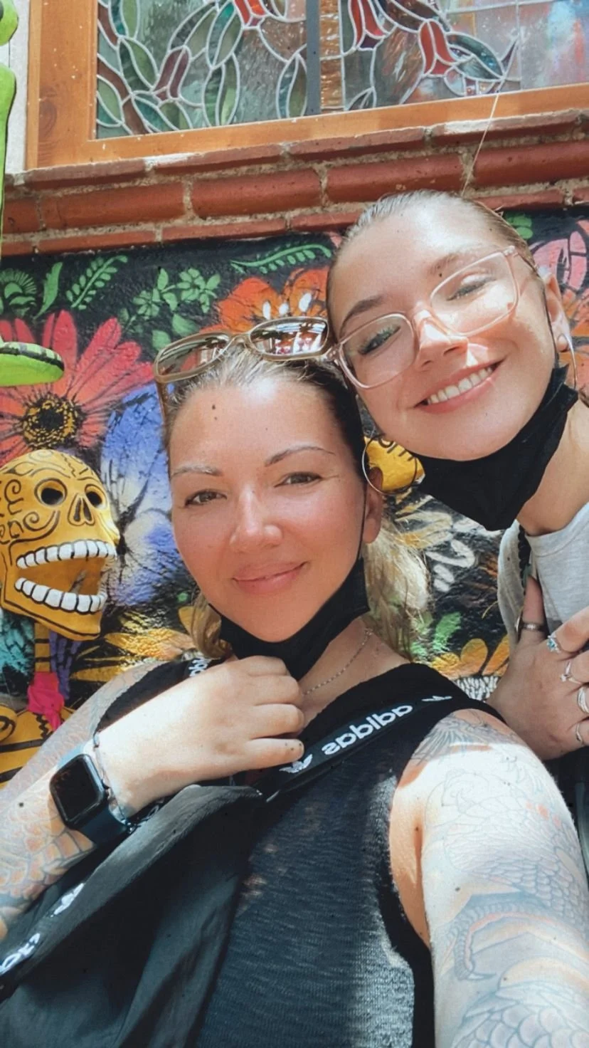 Two smiling women taking a selfie in front of a colorful mural with flowers, a skull, and a bird stained glass window.