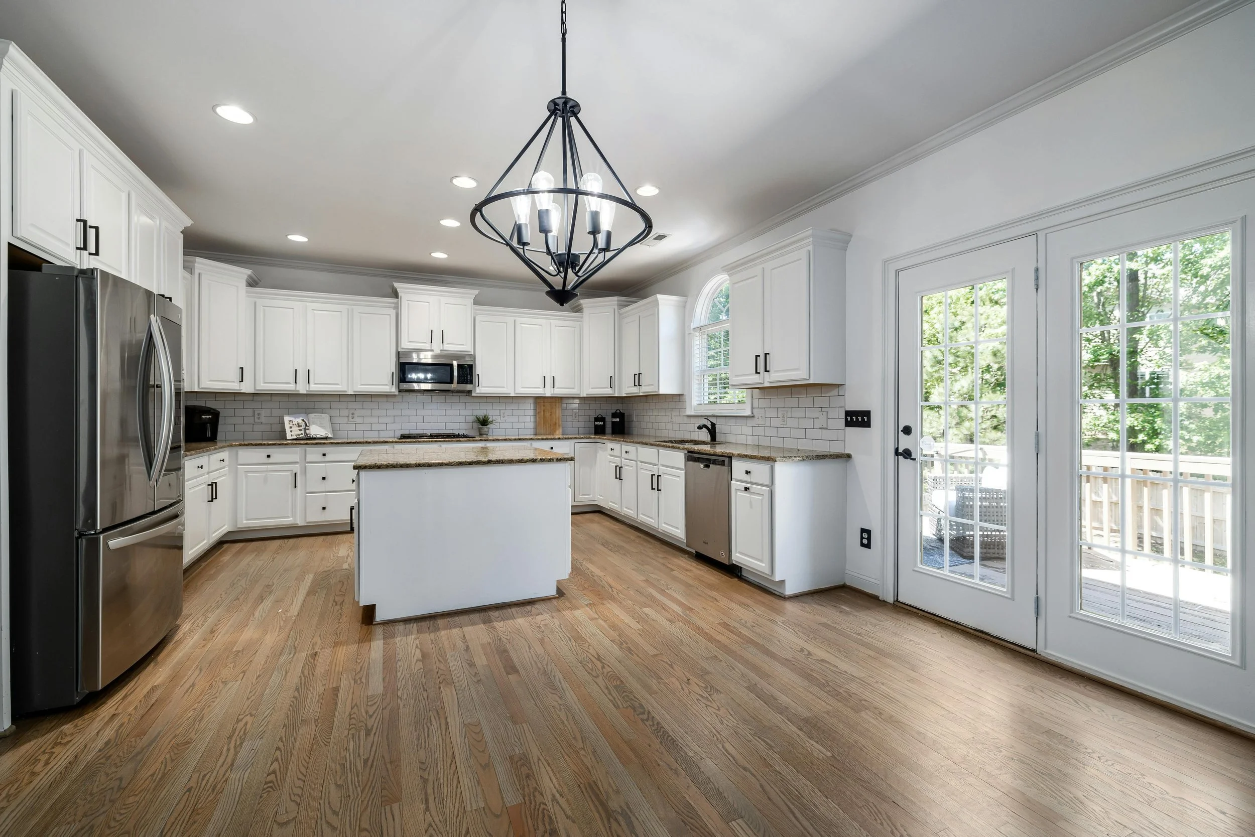 Modern kitchen with white cabinets, granite countertops, stainless steel appliances, hardwood floors, and a chandelier above a central island. Large glass double doors lead to an outdoor deck, with windows showing greenery outside.