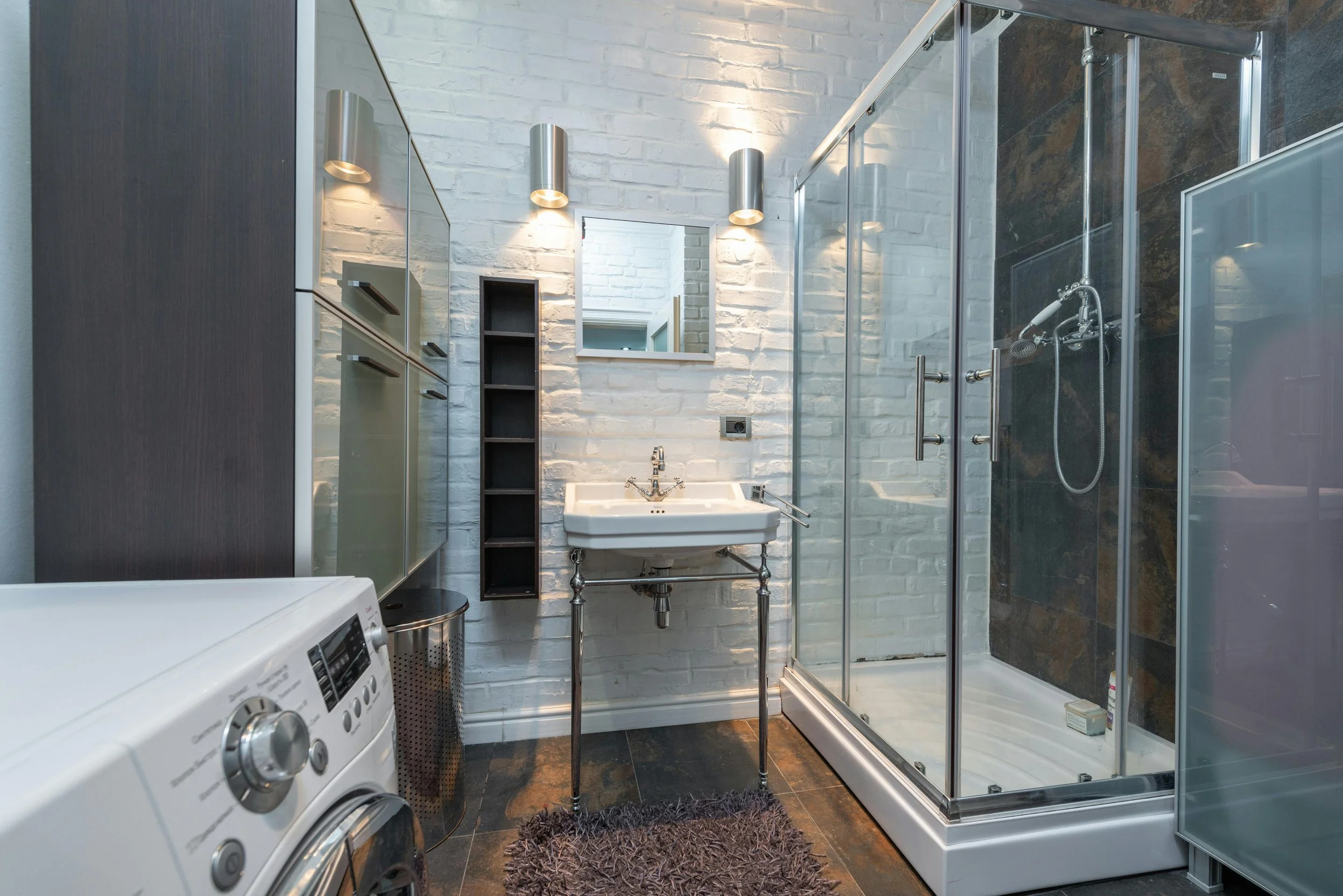 Modern bathroom with white brick wall, shower enclosure, small sink, mirror, and laundry washing machine.