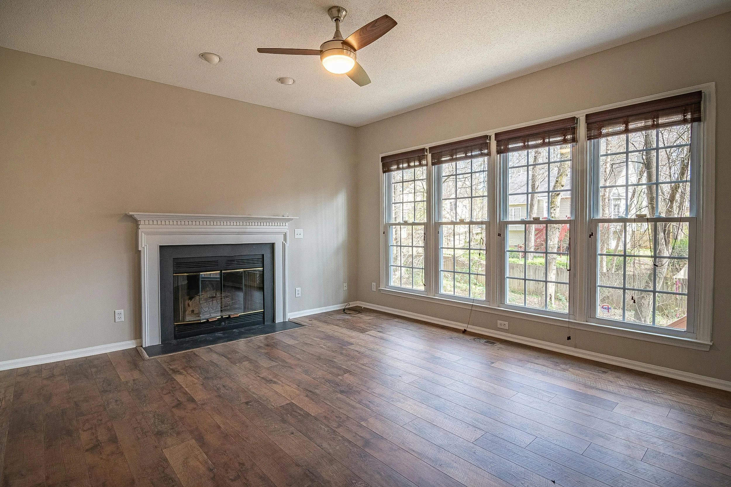 Empty living room with large windows, fireplace, wooden floor, ceiling fan, beige walls, and window blinds.