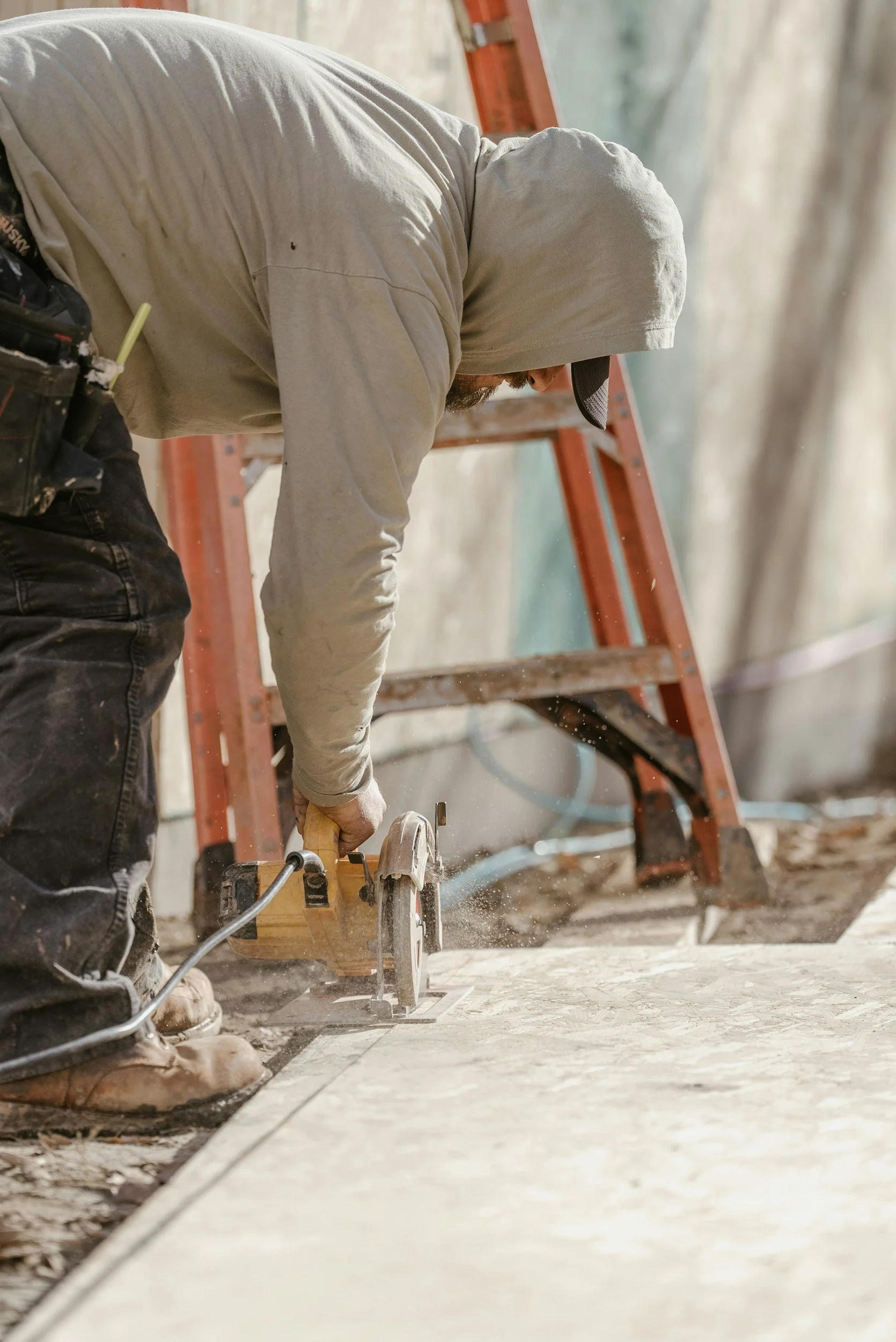 A worker in a hoodie and work boots cuts a concrete slab with a circular saw at a construction site, with a ladder and tarp in the background.