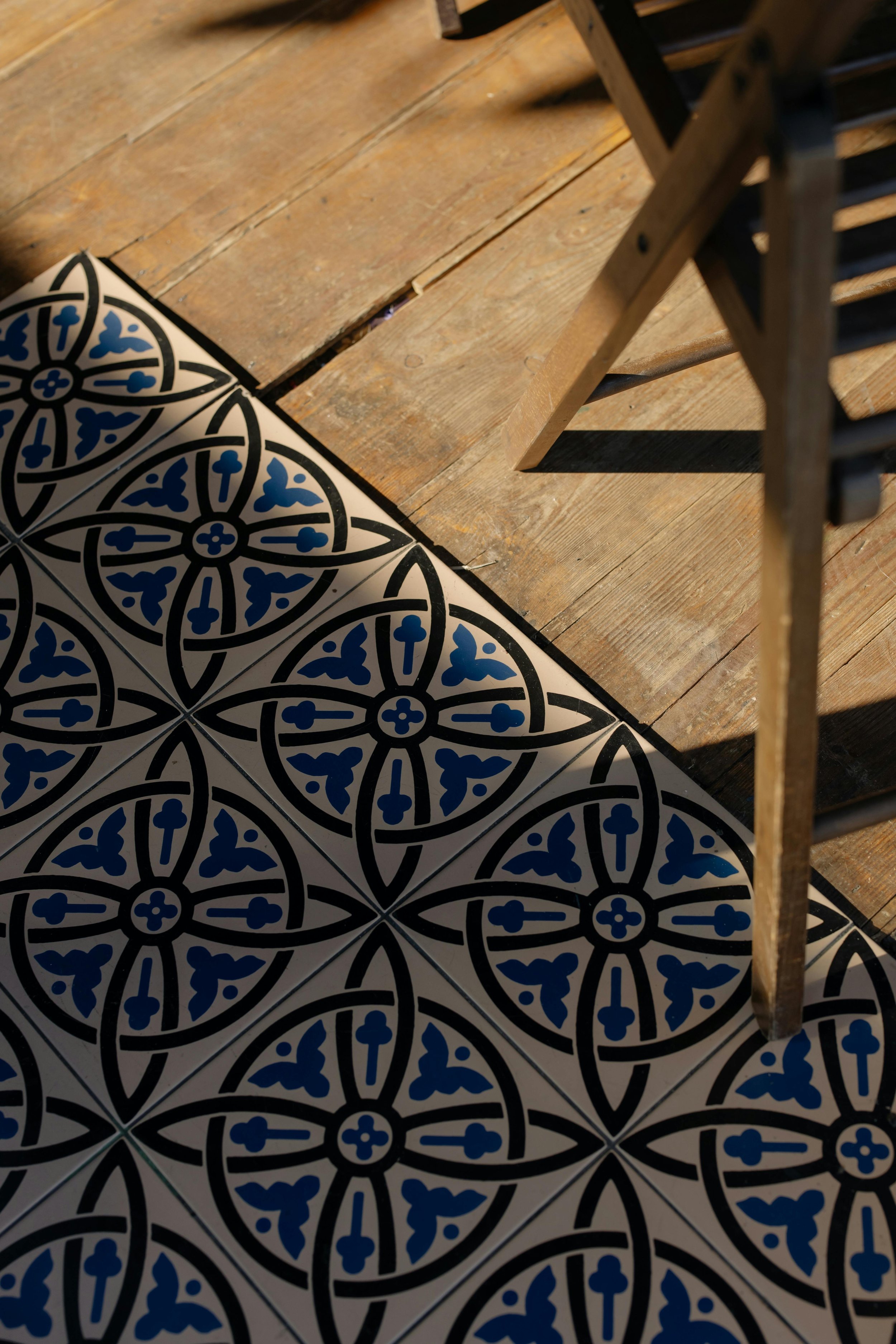 Close-up of a patterned tiled floor with black and blue designs and a wooden chair on a wooden floor with sunlight creating a shadow.