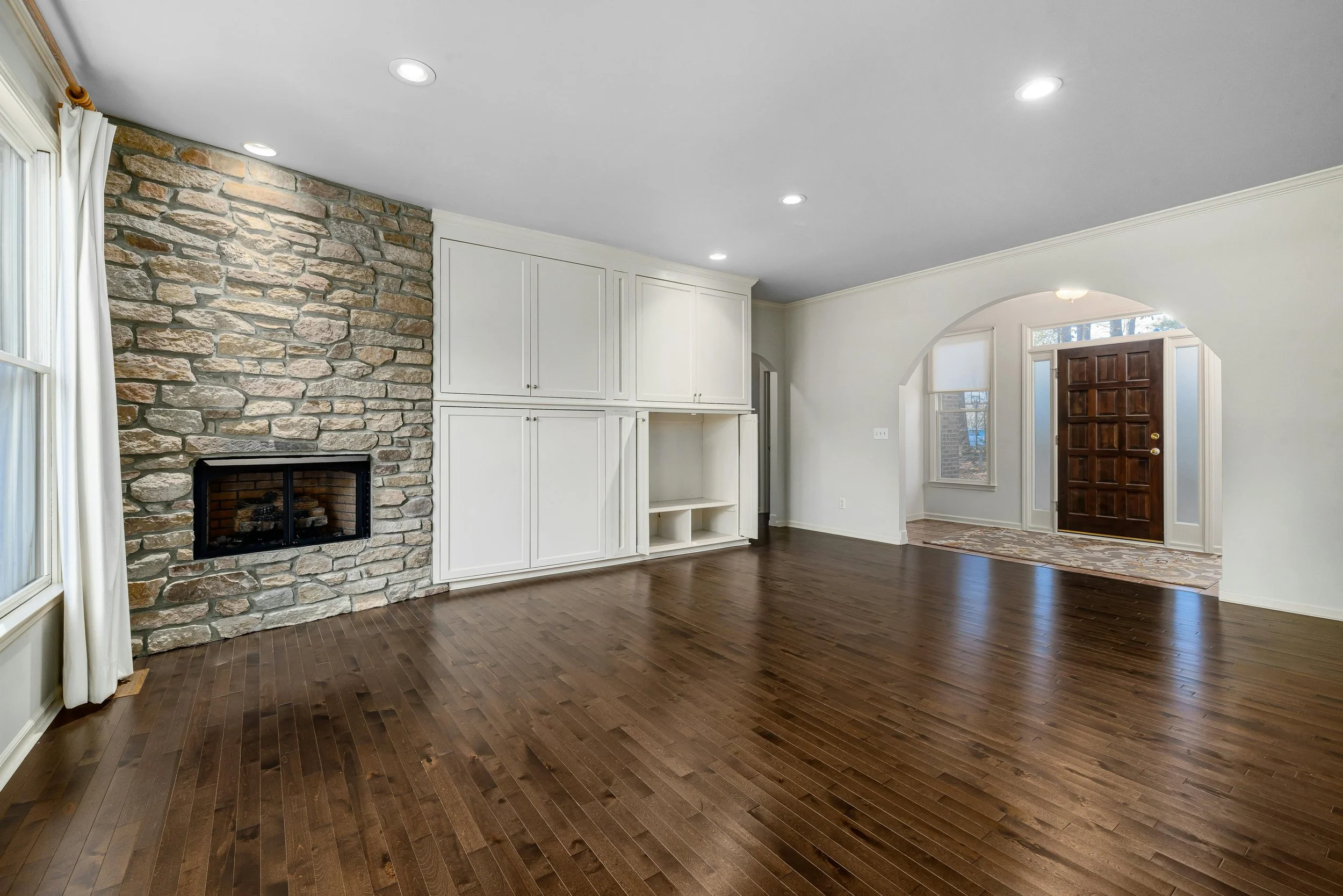 Empty living room with a stone fireplace, white built-in cabinets, hardwood floors, and a front door with an archway leading to the foyer.
