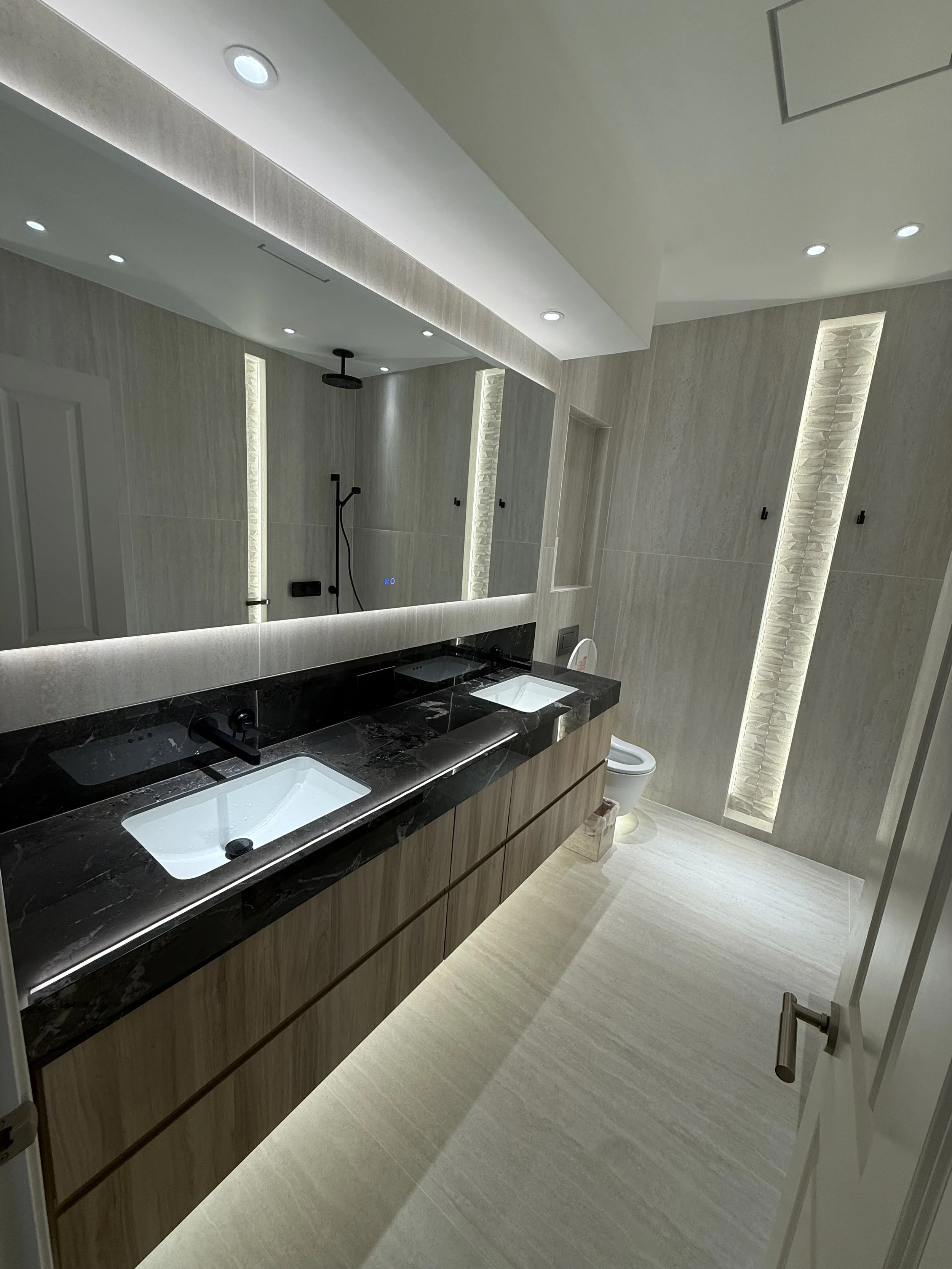 Modern bathroom with double sink vanity, black marble countertops, large mirror, beige textured walls, lit vertical niches, toilet, and walk-in shower with black fixtures.