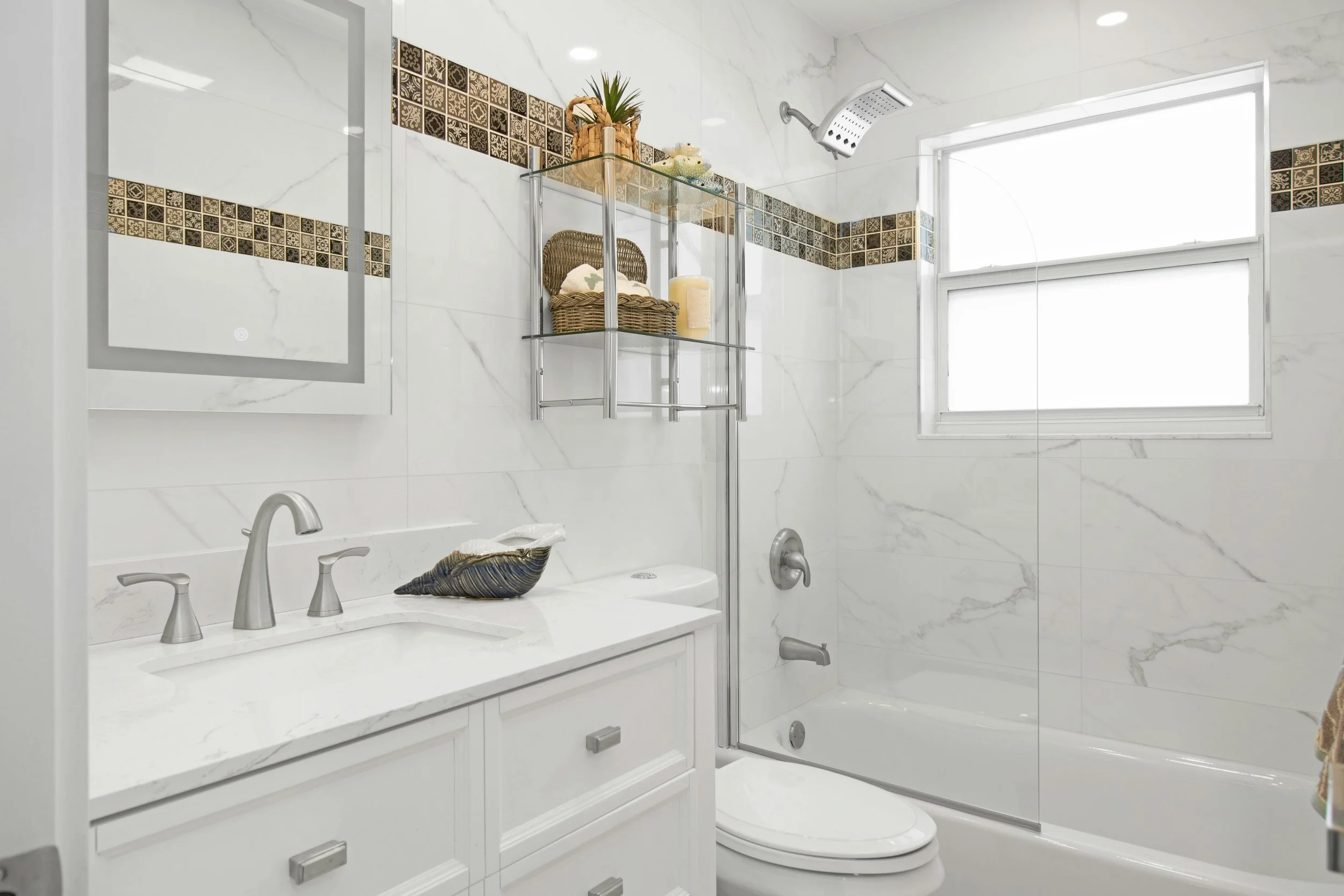 Clean bathroom with white marble walls, a bathtub with a showerhead, a window, a white vanity with a sink, and decorative baskets on a shelf.
