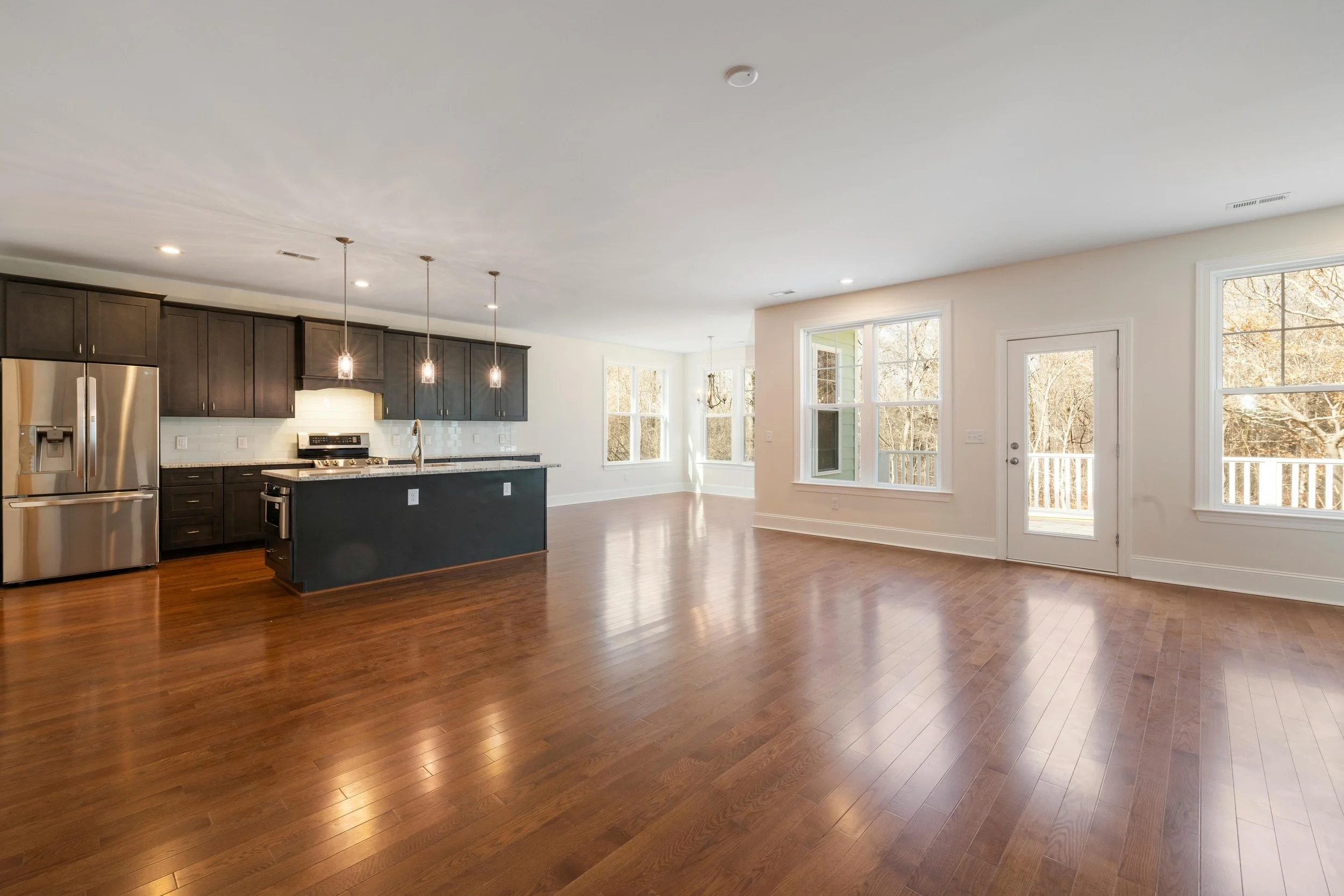 Open-concept living space with hardwood floors, kitchen with dark cabinets, stainless steel refrigerator, island with pendant lighting, and large windows with a view of trees outside.