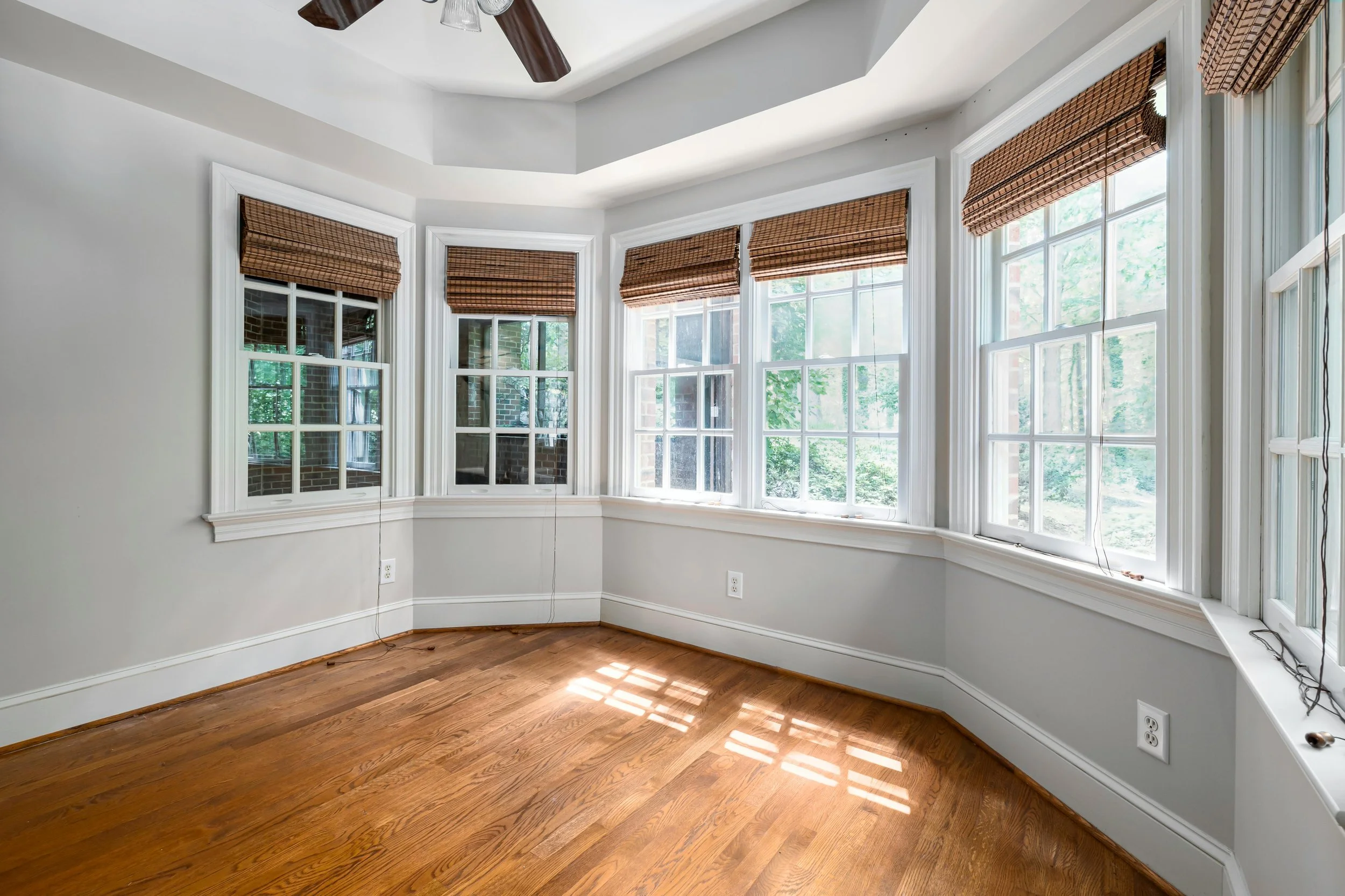 Empty room with hardwood floors, white walls, multiple large windows with brown woven blinds, and a ceiling fan.