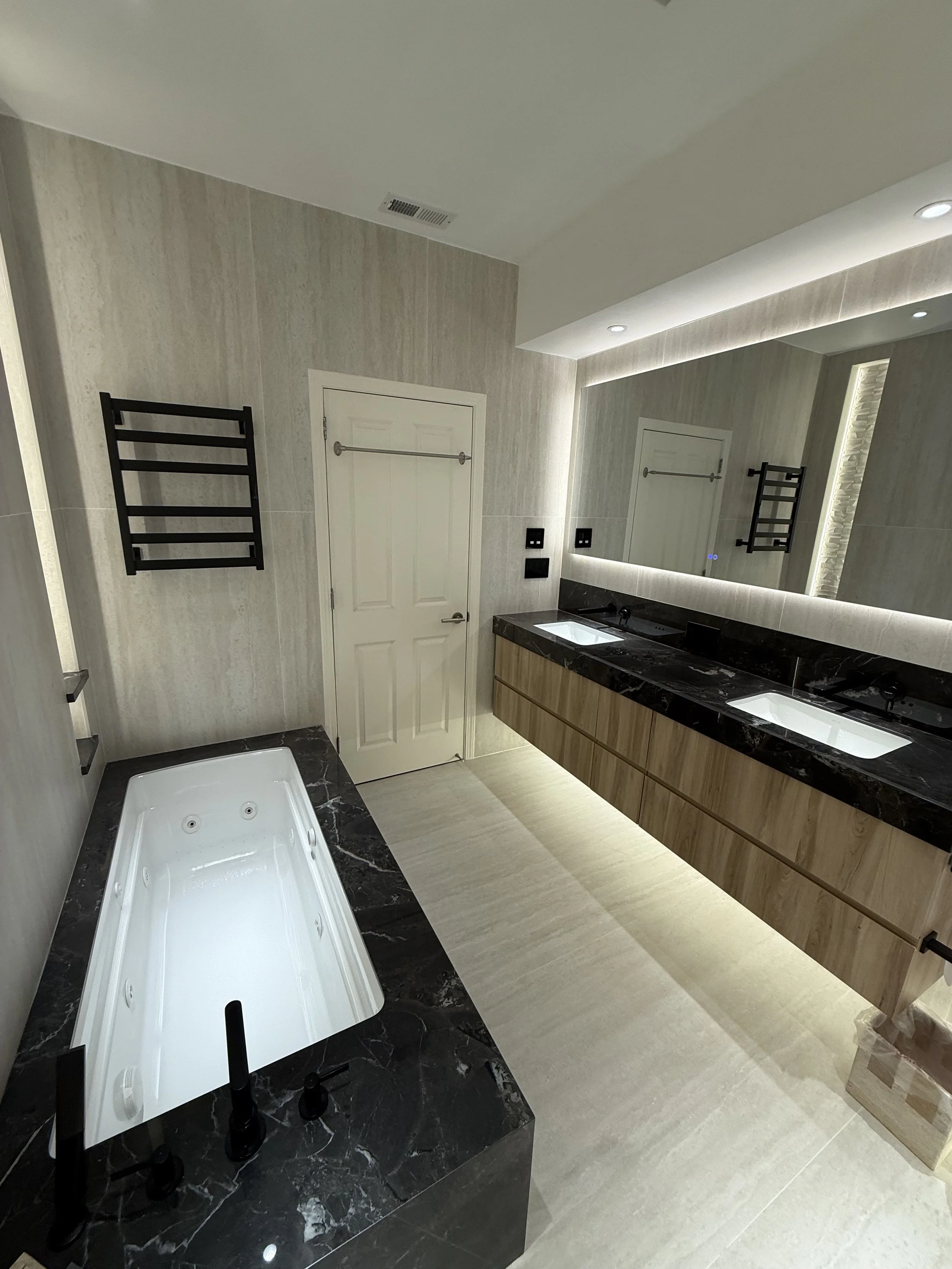 Modern bathroom with a built-in bathtub with black marble surround, a double vanity with black marble countertop, two sinks, large mirror, and a towel heater on the wall.