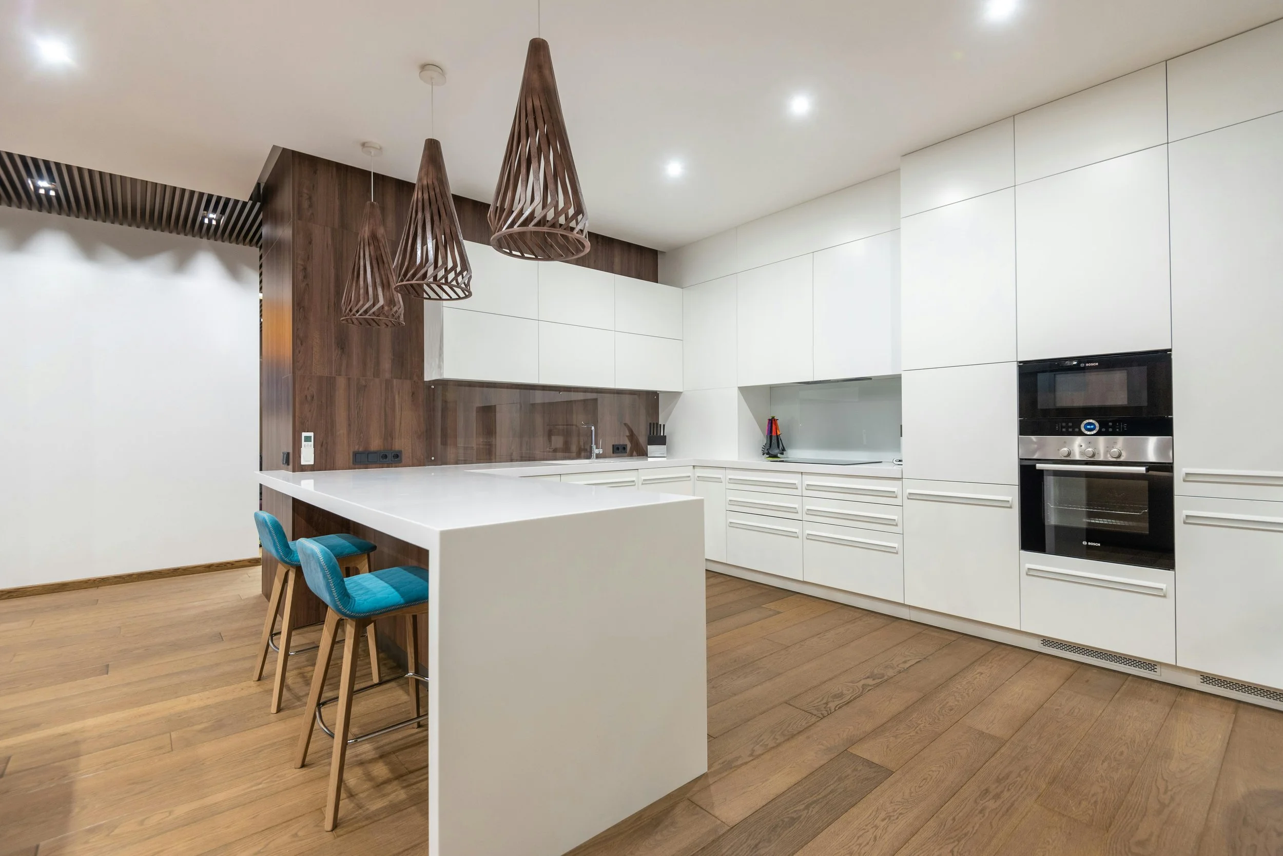 Modern kitchen with white cabinets, wooden accent wall, and blue bar stools at island, featuring three hanging wooden pendant lights.