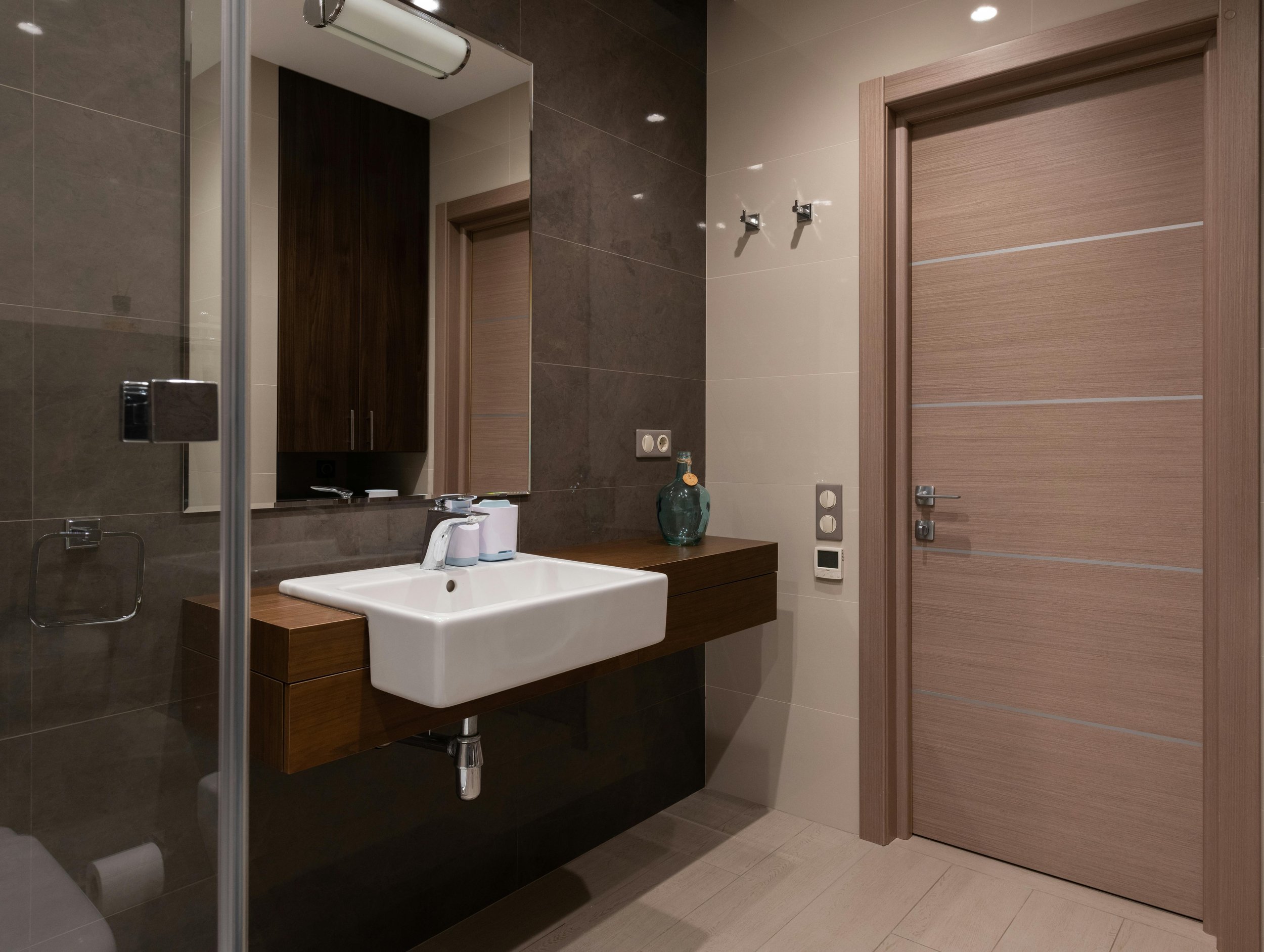Modern bathroom with a wall-mounted sink, a mirror, a wooden cabinet, a door, and various fixtures.