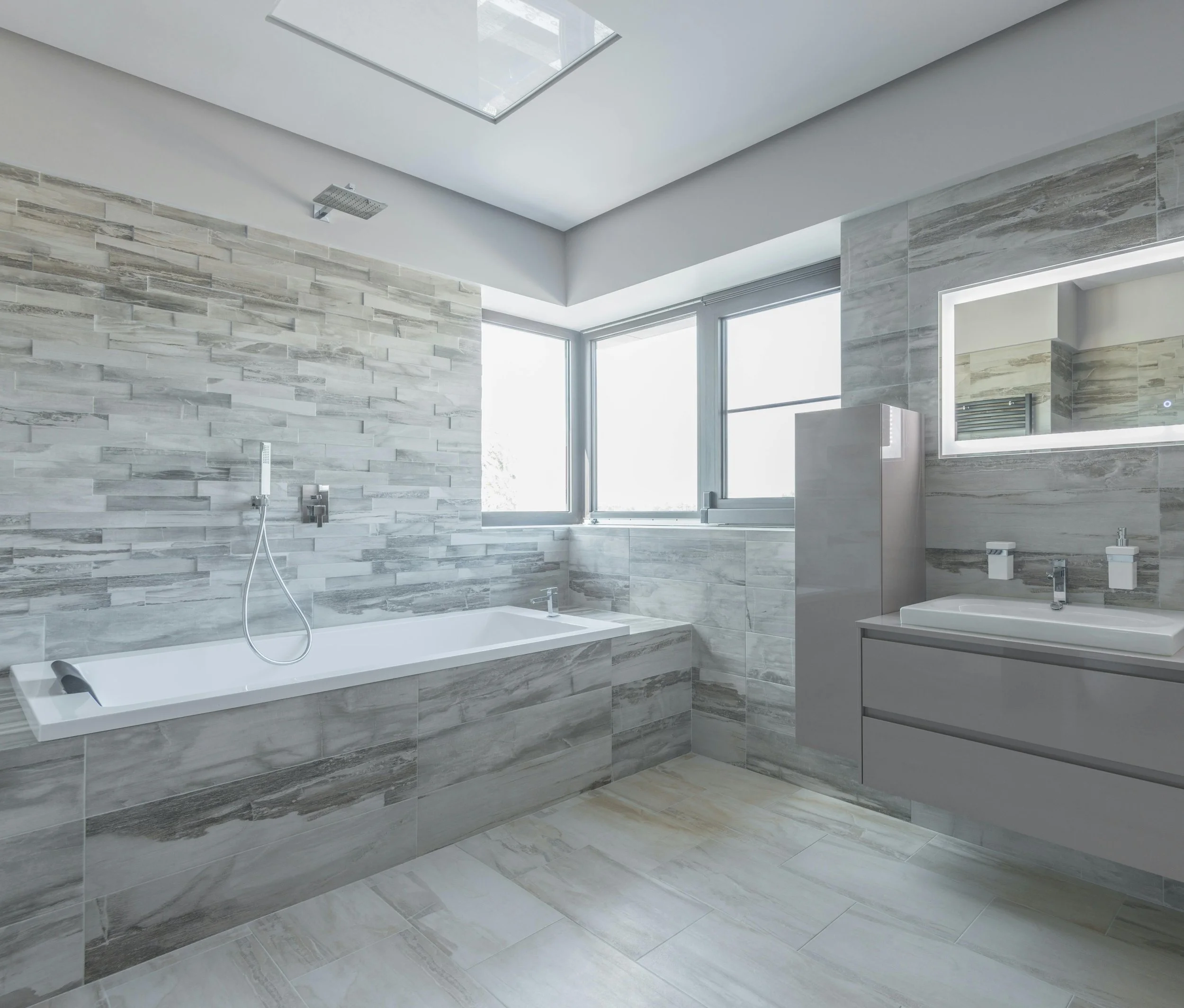 Modern bathroom with gray tiled walls, a built-in bathtub, a large window, a mirror above a sink, and a showerhead on the wall.