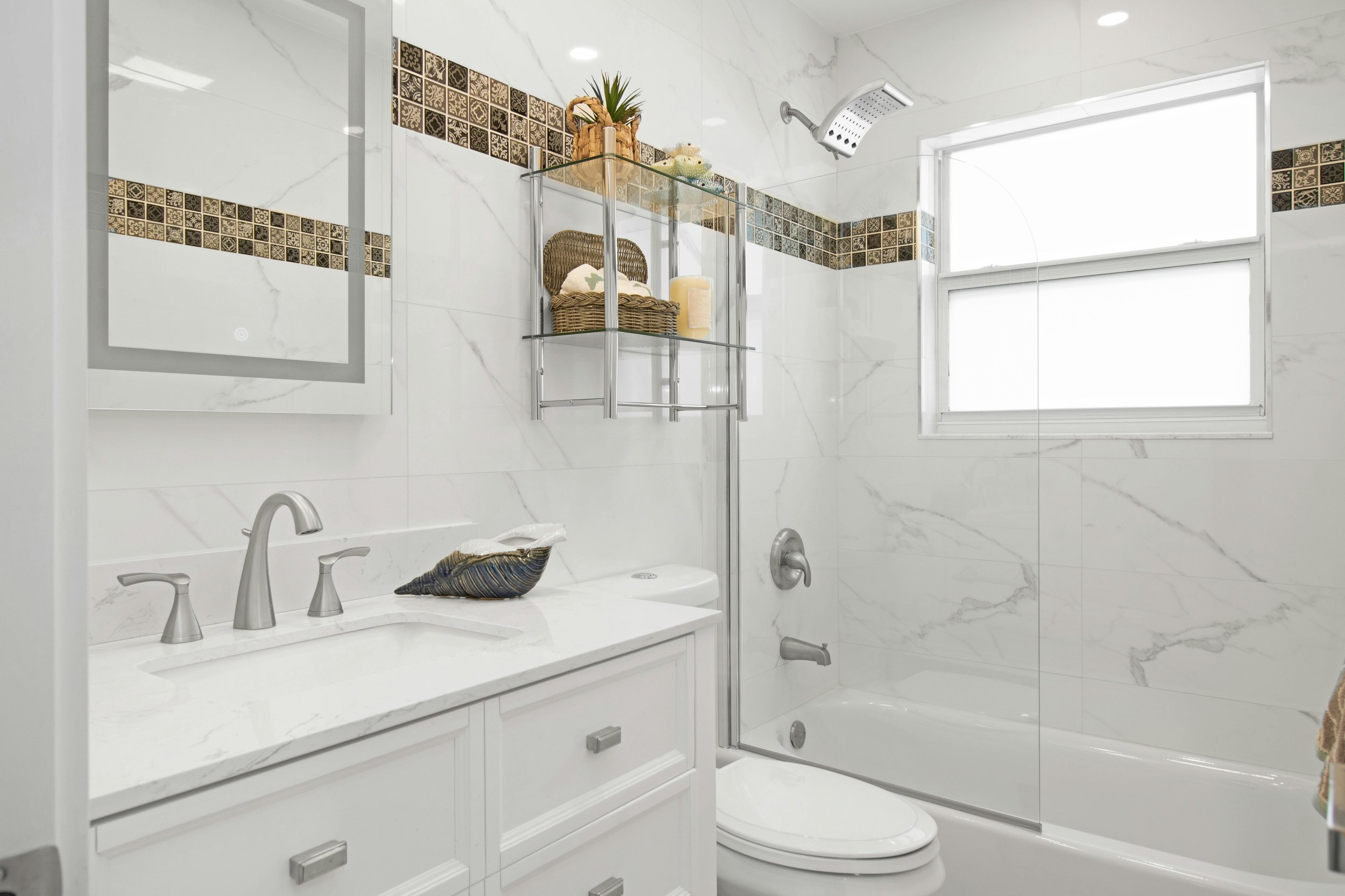 A modern bathroom with white marble walls, a white vanity with a sink and silver fixtures, a toilet, and a bathtub with a glass shower door. There is a window with frosted glass, a wall-mounted shower head, and a metal shelf with decorative baskets, candles, and plants.