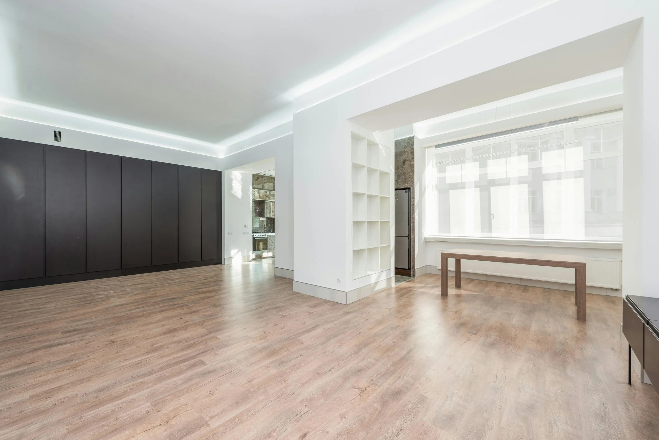 Empty modern apartment living space with large window, wooden floors, and minimal furniture.