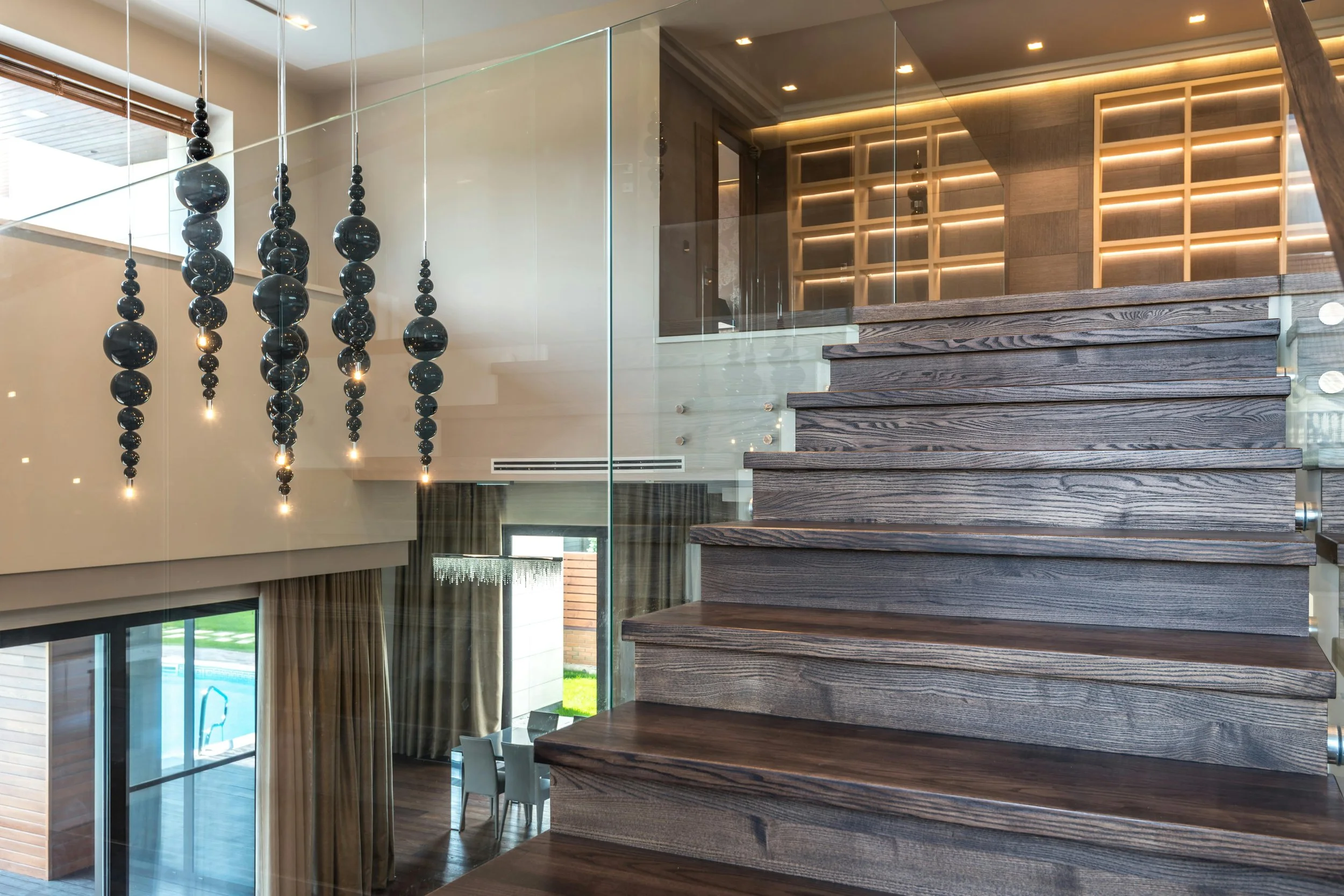 Modern interior with floating dark wood stairs, glass railing, and hanging black glass orb pendant lights in a well-lit space.