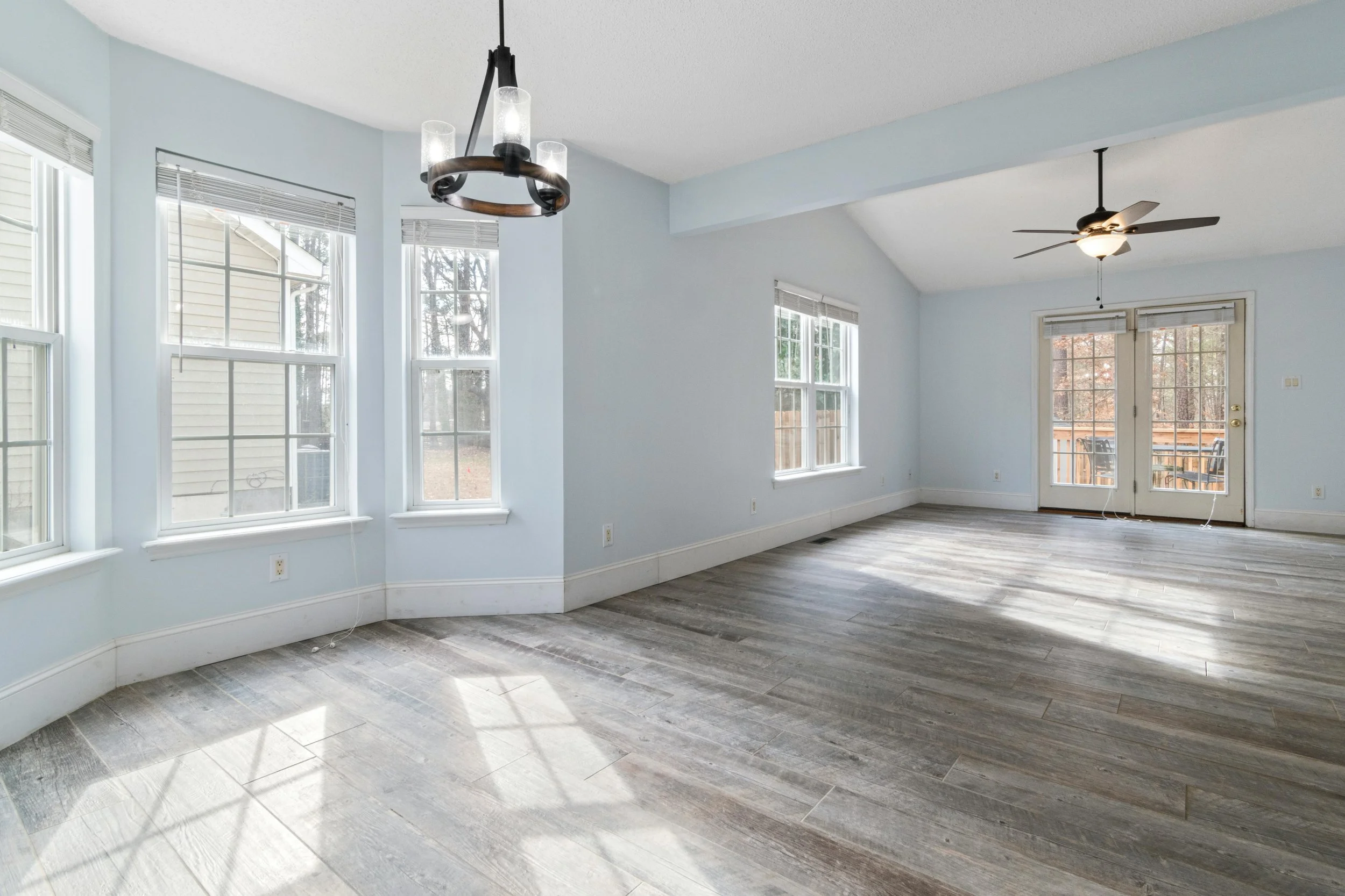 Empty living room with large windows, a ceiling fan, a hanging light fixture, and wood-look flooring.