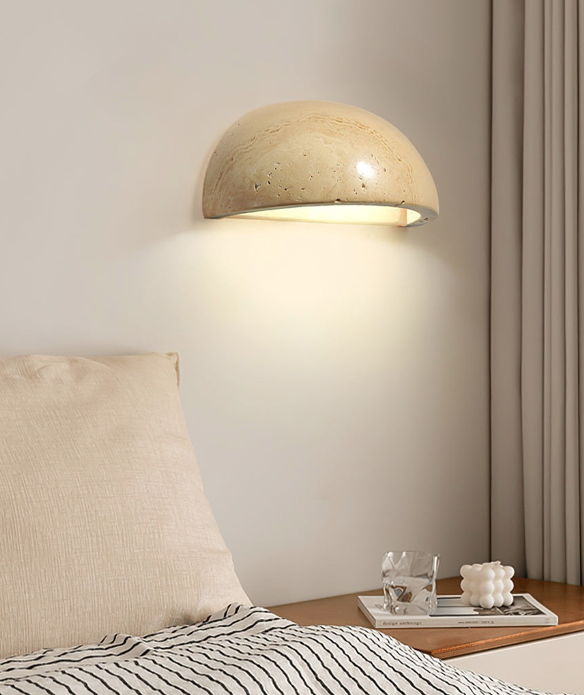 A modern bedroom wall with a half-dome wall sconce emitting warm light, a beige pillow, a striped bedspread, a side table with a glass of water, a white textured candle, and a magazine.