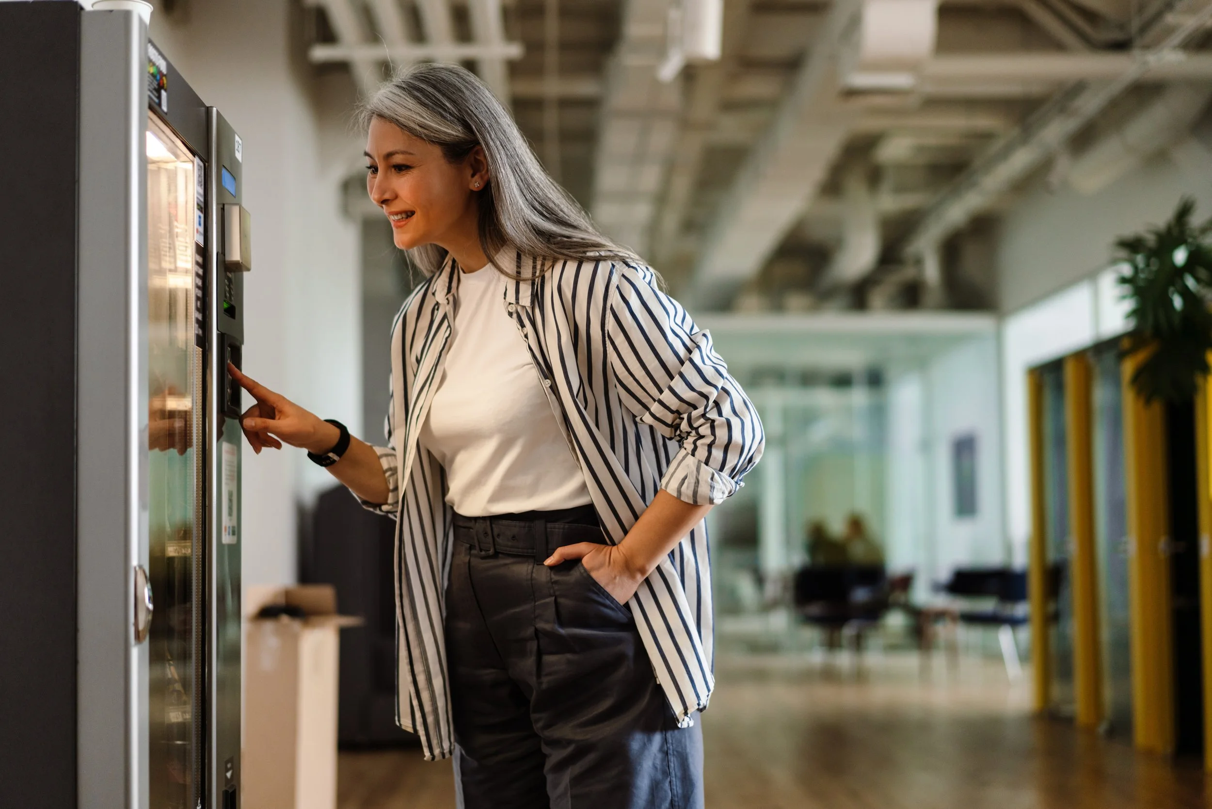 A woman with gray hair wearing a striped shirt and black pants using a vending machine in an office lounge.