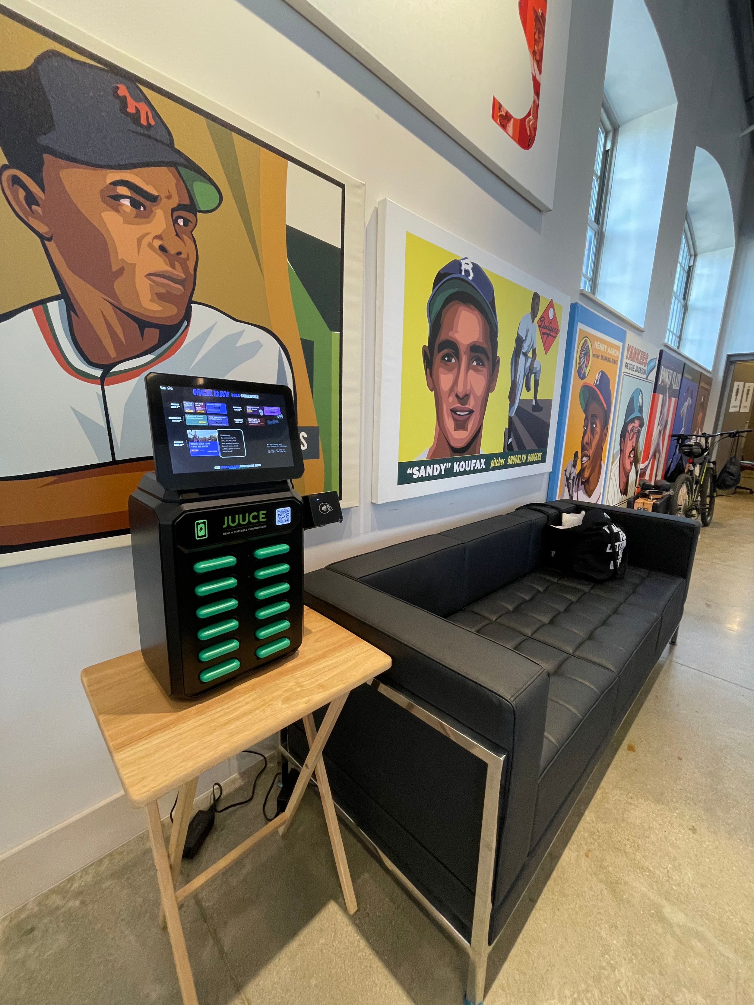 Indoor space with vintage baseball-themed artwork on the wall, a black leather couch, and a wooden side table holding a juice machine with a touchscreen display. Bicycles are visible in the background near large windows.