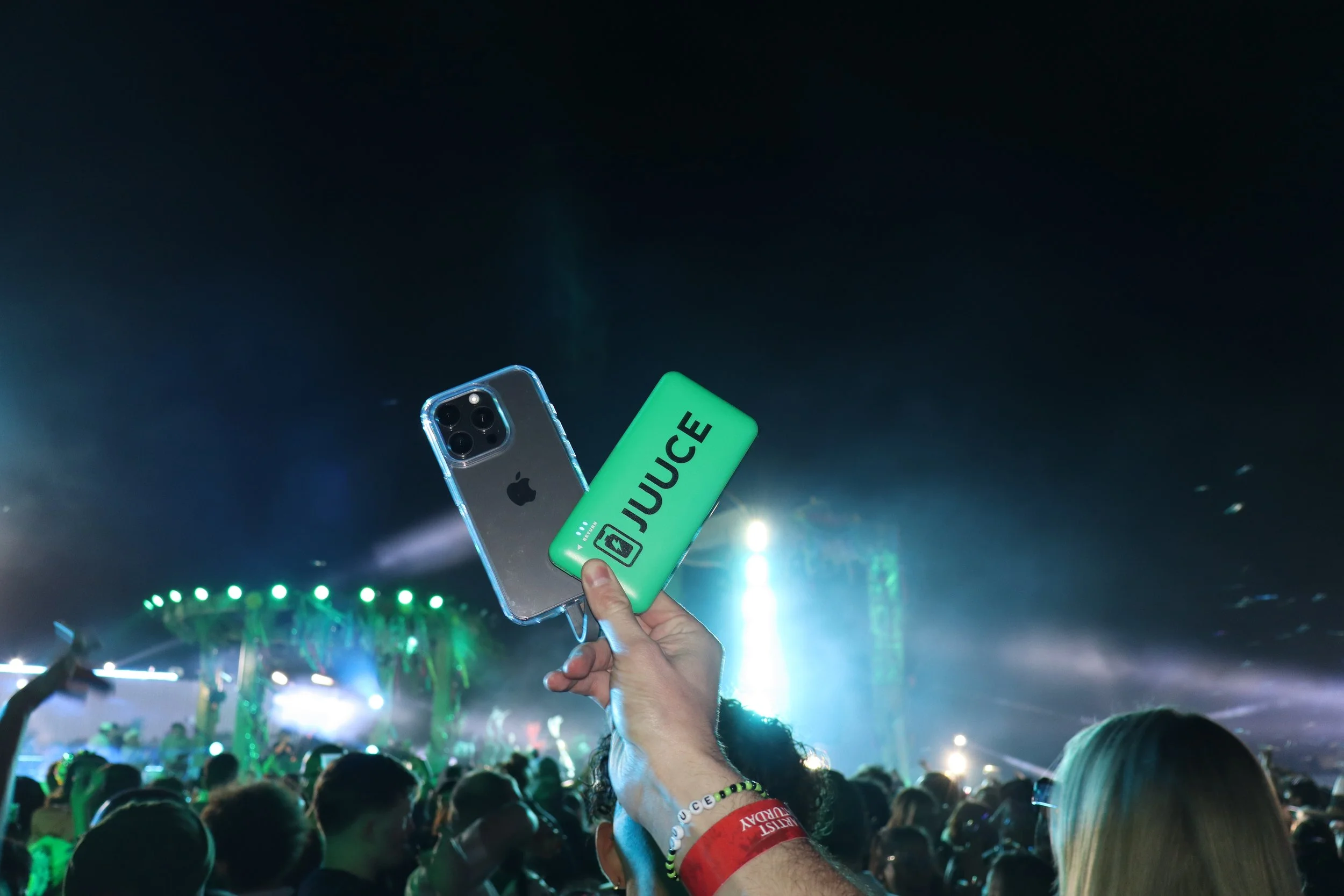 Person holding a green JUICE portable charger and an iPhone at a crowded concert or festival with colorful lights.