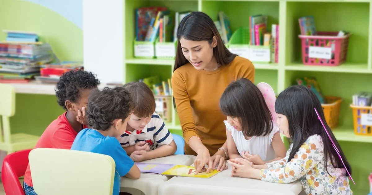 A teacher reading a book to a group of five children in a colorful classroom.