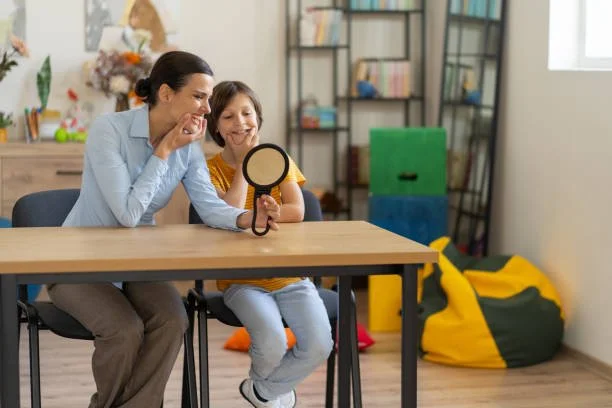 A woman and a girl sitting at a table looking at a mirror together in a room with bookshelves and colorful bean bags in the background.