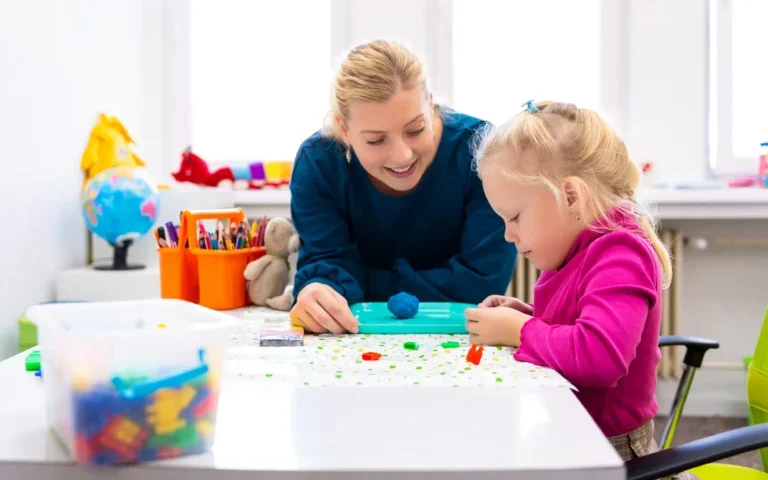 A woman and young girl doing arts and crafts at a table in a classroom setting.