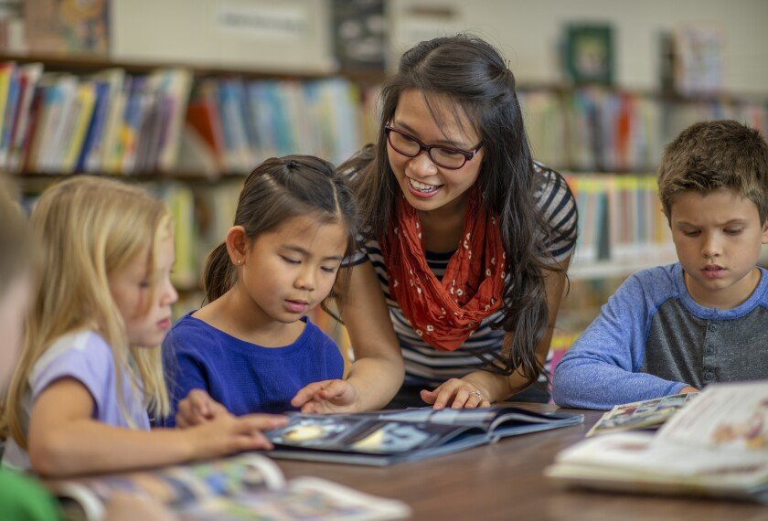 A teacher with glasses and a red scarf reading a book with three children in a library.