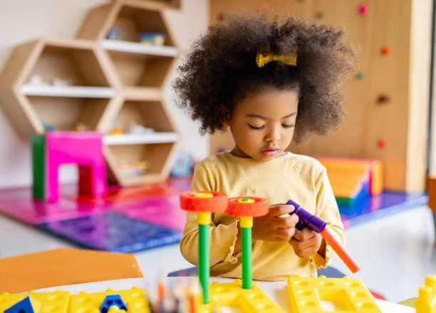 Young girl with curly hair and a yellow shirt playing with colorful building toys in a playroom.