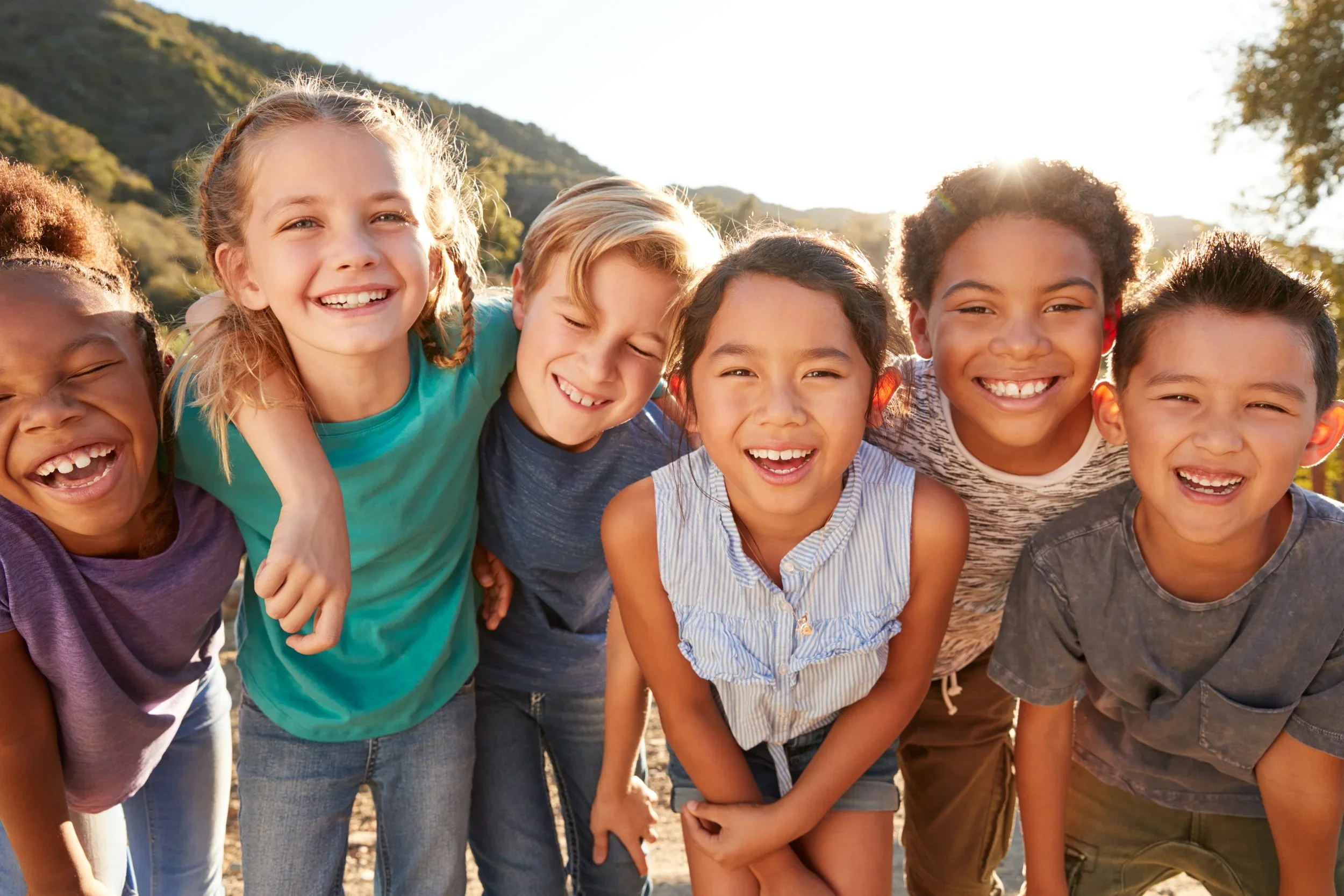 Group of seven diverse children smiling and laughing outdoors during daytime with mountains and trees in the background.